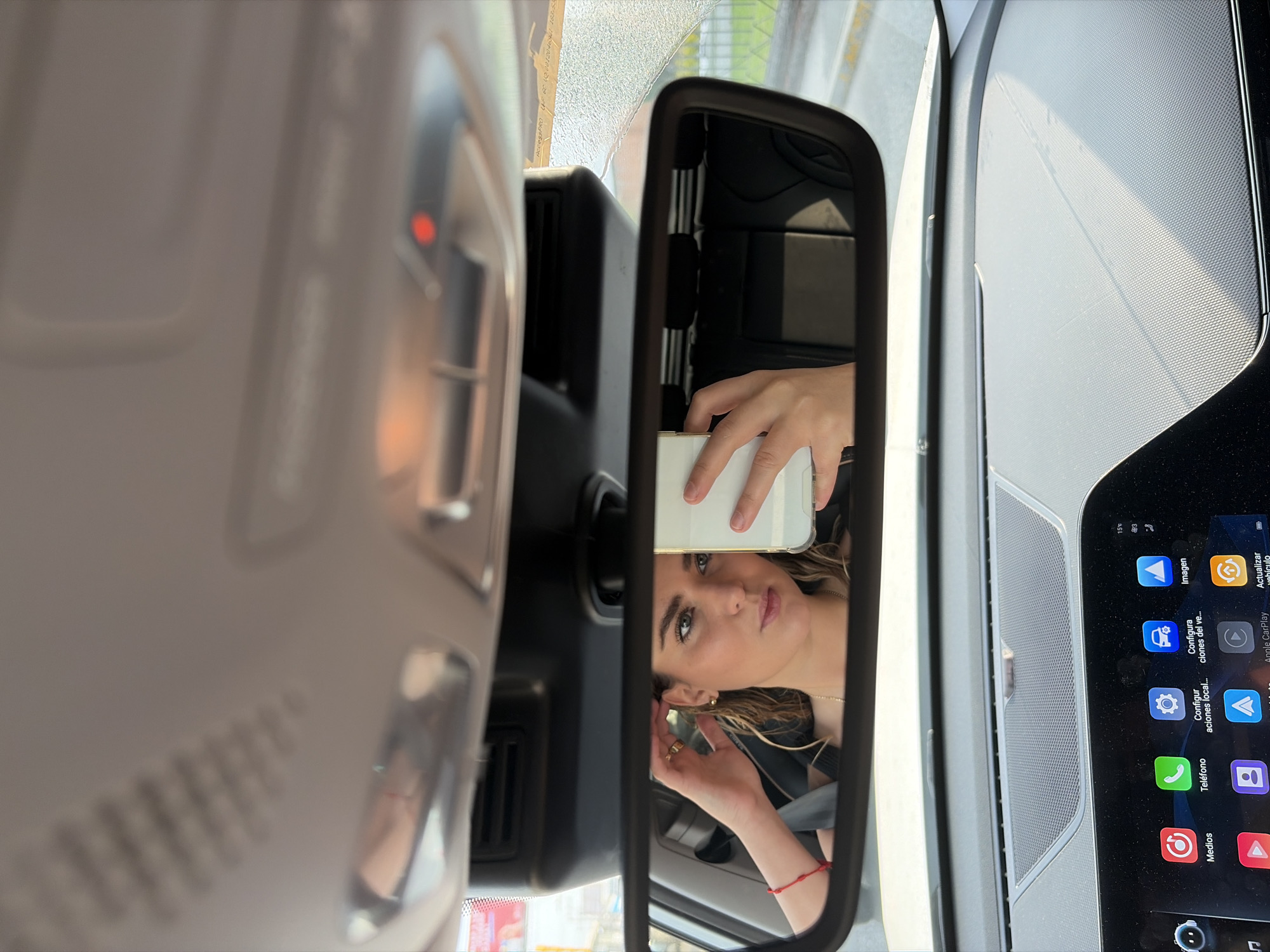 This is an interior shot taken inside a car. The focal point is the rearview mirror, in which a woman is taking a selfie with her smartphone. She is fair-skinned, with light brown hair, and is wearing a black jacket and a gold necklace. Her left wrist is adorned with a red bracelet.
To the left of the mirror is the car's interior ceiling, which is light grey and features a control panel with a red light. To the right of the mirror is the dashboard, which includes a large infotainment screen displaying various application icons in Spanish, such as "Imagen" (Image), "Teléfono" (Telephone), and "Medios" (Media). The screen indicates a temperature of 15°C.
The OCR text from the infotainment screen includes technical codes related to vehicle configuration and updates, suggesting the car is equipped with advanced tech features. The text also notes "AccessPRO LUF EC VEDO. 001204641 183" on the windshield.
The image appears to be taken in San Bernardino Tlaxcalancingo, Mexico, given the information on the windshield.