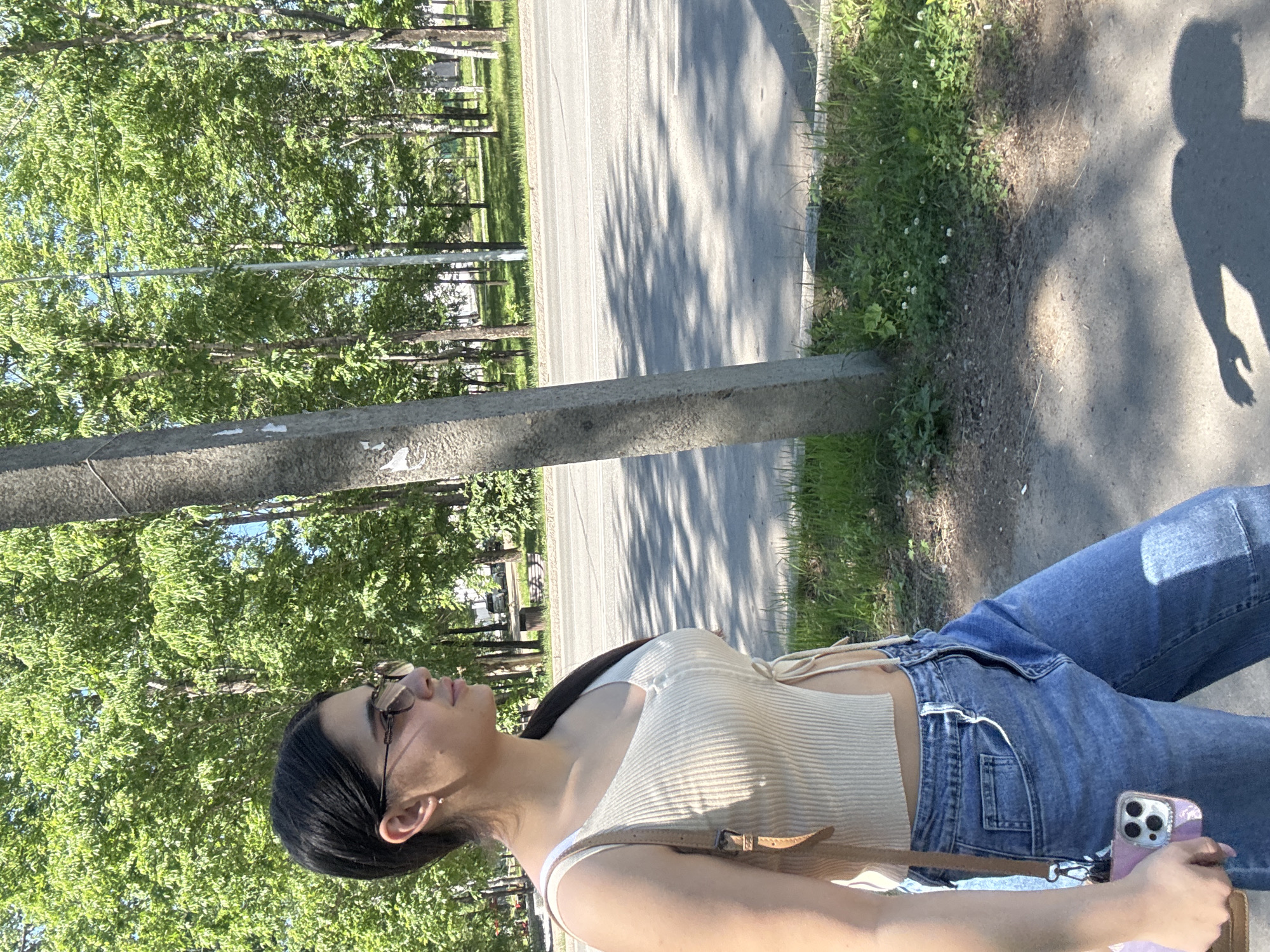 A young woman with dark hair and sunglasses is walking on a sidewalk next to a road in Birobidzhan, Russia. She is wearing a cream-colored, ribbed tank top and blue jeans. She is carrying a light brown purse over her shoulder and holding a pink phone in her left hand. The background consists of trees with green leaves, a street, and a sidewalk. The sun is shining, casting shadows on the ground.
