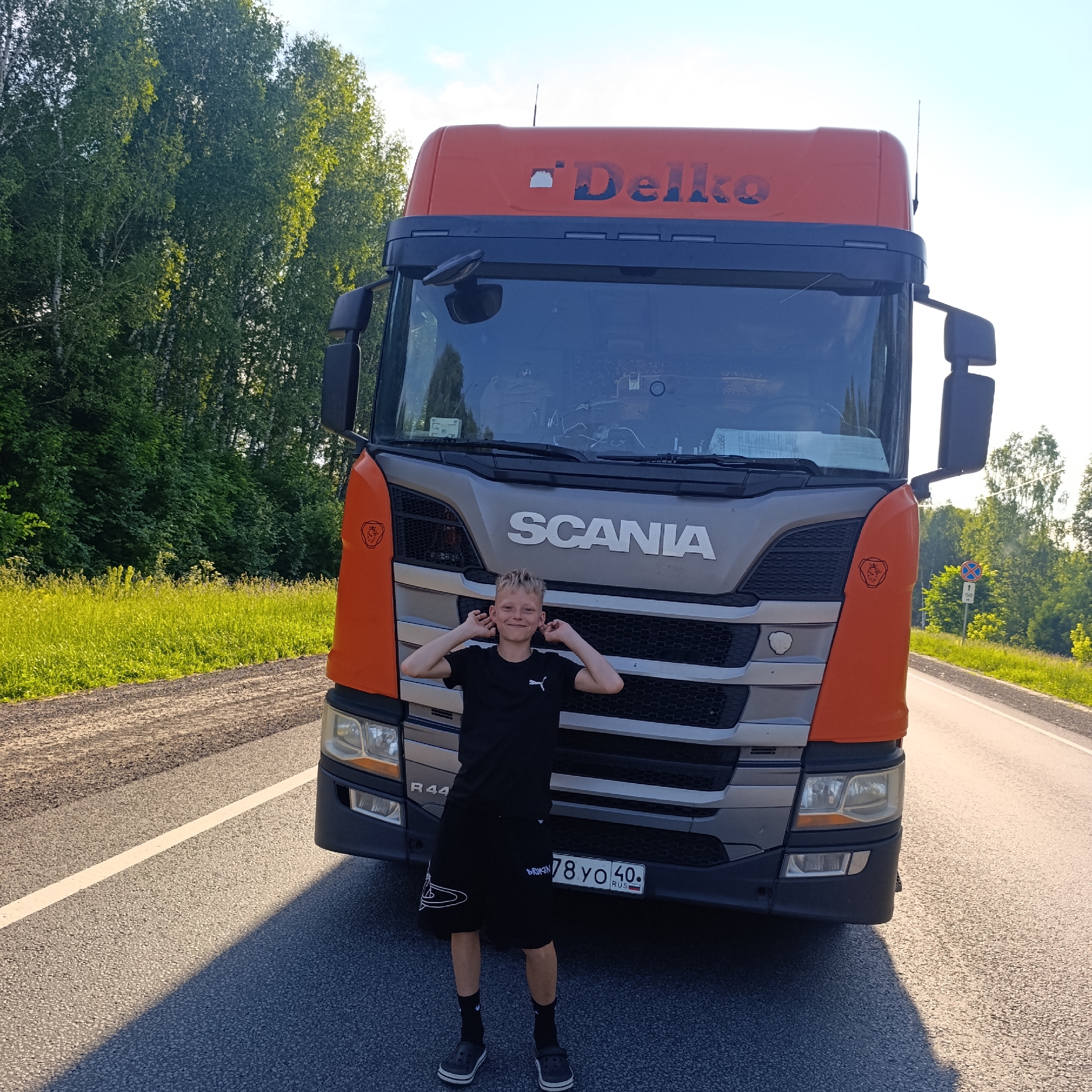 A young boy with blond hair, wearing a black t-shirt and black shorts, stands in front of a large orange and silver Scania truck on a paved road. The boy has his hands raised to his ears and is smiling at the camera. The truck has the word "SCANIA" prominently displayed on its front grille. The truck's upper section is orange with the logo "Delko" on it. The background features a line of trees and a bright sky. The scene appears to be outdoors, possibly on the outskirts of a city like L'vovo, Russia, given the presence of trees and a road. A Russian license plate on the truck reads "78 YO 40".
