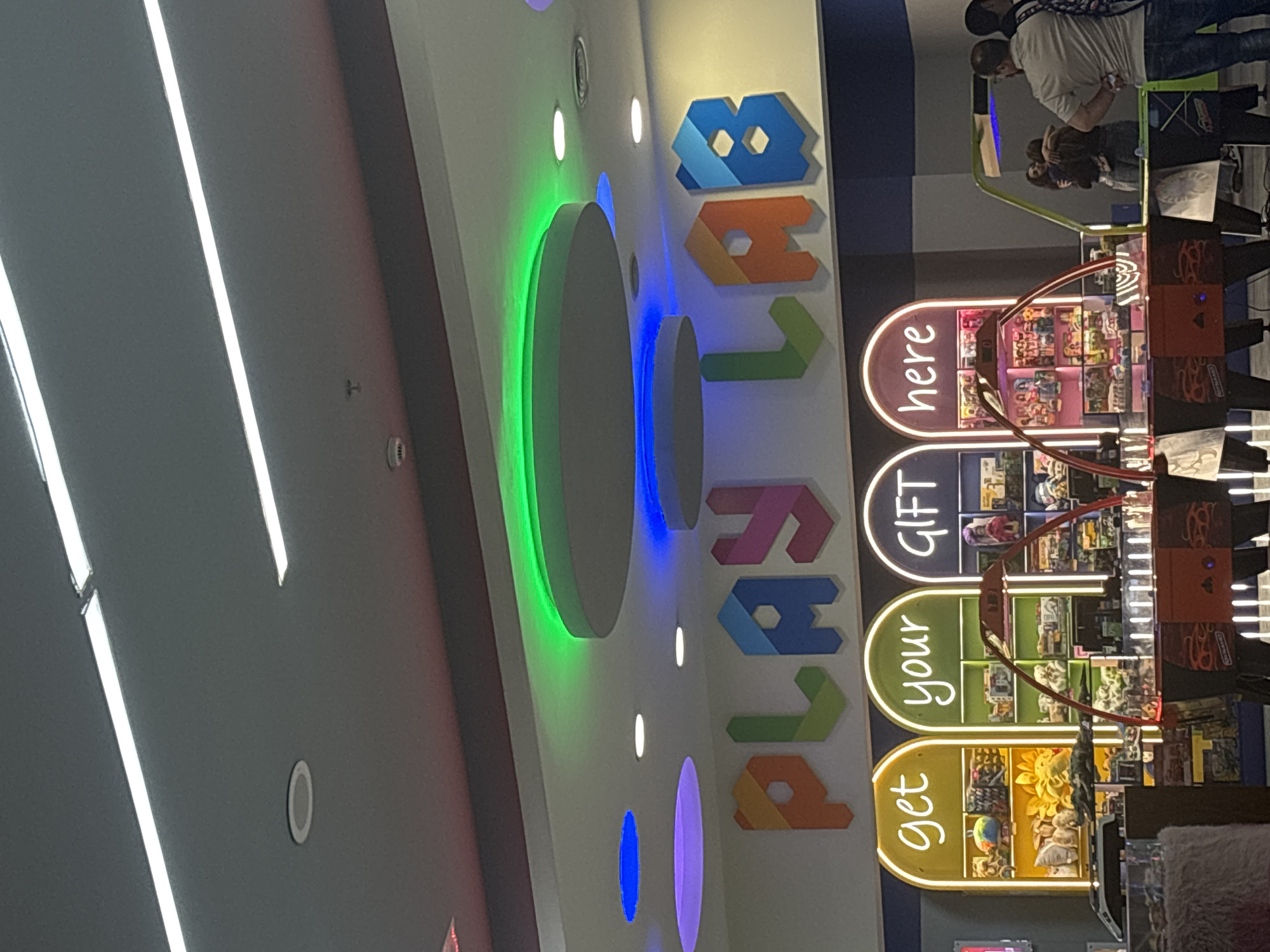The image captures an indoor entertainment or arcade area, likely in Yerevan, Armenia, characterized by vibrant lighting and a modern, playful design.
A prominent feature is a large, vertically oriented sign on the left side of the frame, spelling "PLAY LAB" in colorful, block-shaped letters. The letters are rendered in various hues, including blue, orange, and green. Below this sign, a series of arcade machines, possibly claw machines, line the right side of the space. These machines feature reddish-orange casings with decorative patterns and brightly lit display sections showcasing numerous toys and prizes. Integrated into these machines are arched, illuminated display panels with text labels: "get" on a yellow background, "your" on a green background, and "GIFT here" on a pink/purple background, suggesting a prize redemption area.
Two adults are visible standing behind these arcade machines, reflected in their glass panels. One person wears a light-colored top and a backpack, seemingly observing the games or activity.
The lighting throughout the space contributes significantly to the atmosphere. Long, linear fluorescent or LED lights run across the ceiling. On the wall behind the "PLAY LAB" sign, several circular fixtures emit soft, ambient light in green, blue, and purple tones, interspersed with smaller white circular lights, creating a dynamic and engaging environment. The overall scene suggests a lively recreational setting.
