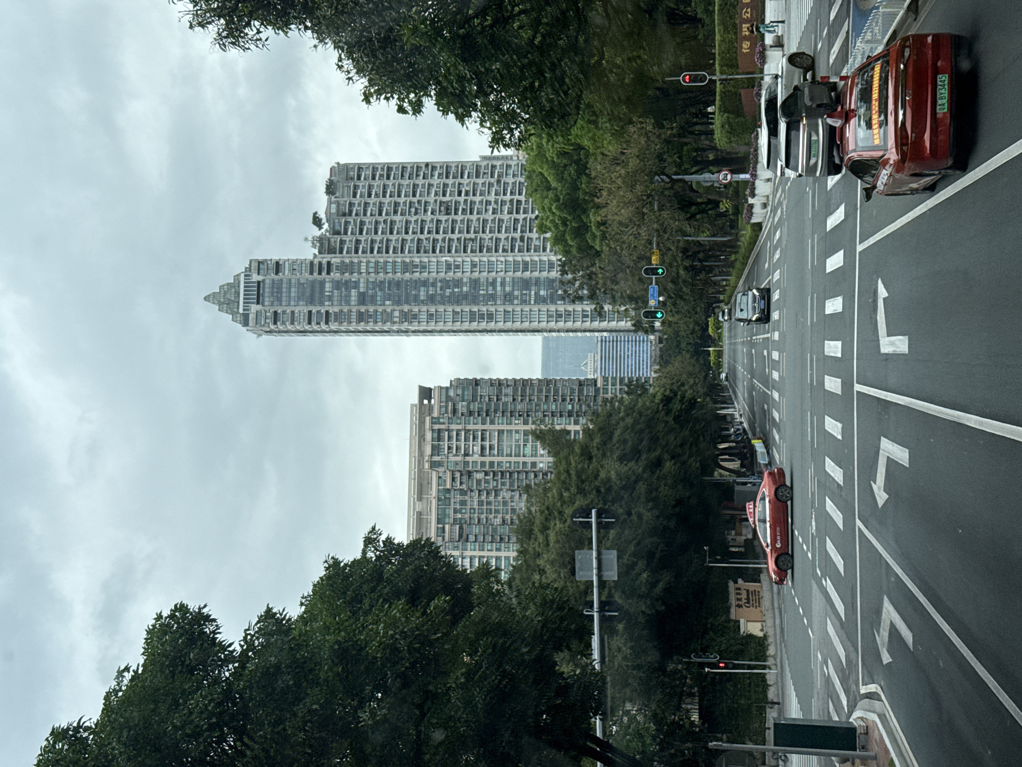 The image depicts a street in Yuexiu District, China, lined with trees and tall buildings. The street has multiple lanes marked with white arrows and pedestrian crosswalks. Several cars are visible on the street, including a red taxi. Traffic lights are present, with some showing red lights. The sky is overcast.