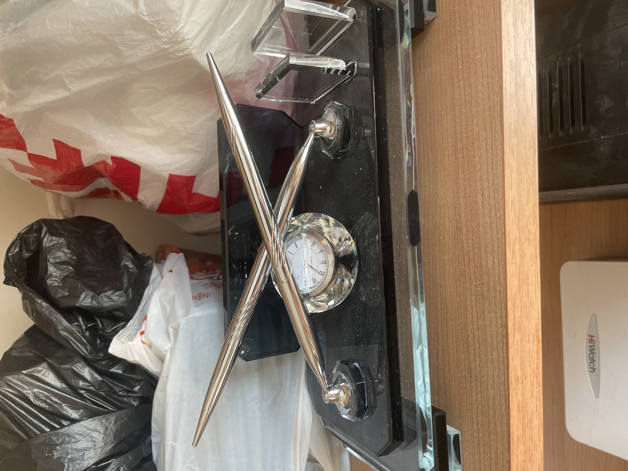 The image displays an indoor scene, likely on a shelf or desk, in Ihtiman, Bulgaria. The central focus is a decorative desk set featuring a small, round clock and two sleek, silver-colored pens crossed in an 'X' formation.
The clock has a white face adorned with black Roman numerals and the word "QUARTZ." Its hands indicate approximately 1:55. The clock face is set within a clear, faceted crystal or glass surround, which reflects light. The pens are pointed and made of a shiny metallic material. The base of the desk set is a black, possibly polished stone or acrylic material, with clear glass or acrylic decorative elements mounted above and below the pens. The surface of the black base appears dusty.
In the background to the left, several plastic bags are visible. A large, crumpled white plastic bag with indistinct red patterns or markings covers much of this area. Partially visible underneath and beside it are black plastic bags, suggesting a cluttered storage area.
To the right of the desk set, a light brown wooden surface, possibly part of a cabinet or shelving unit, is present. Further to the right and partially obscured by the wooden frame, a white rectangular electronic device can be seen. This device features the brand name "HiWatch" written in red and black lettering.
No people or discernible activity are present in the static image. The lighting appears to be ambient indoor light.