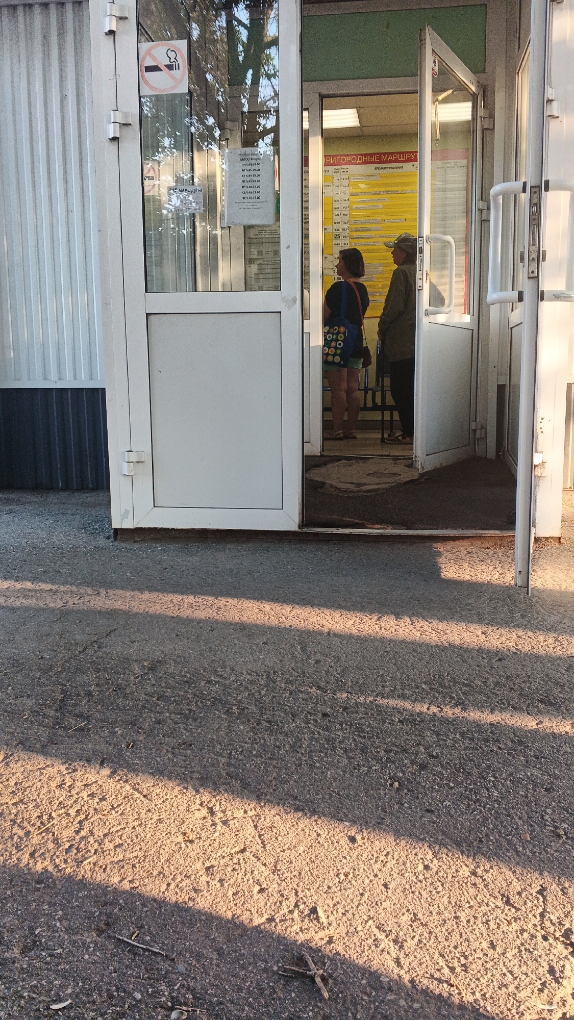 The image shows the entrance to a bus station building in Izhevsk, Russia. The building is white with glass doors and windows. Inside the building, there are two people standing, a woman and a man. The woman is wearing a black shirt and shorts, and is holding a colorful bag. The man is wearing a green jacket and a hat. There is a timetable on the wall inside the building. The ground outside the building is asphalt, and there are shadows from trees on the ground. There is a sign on the door of the building with a cigarette symbol, indicating it is a no-smoking area.