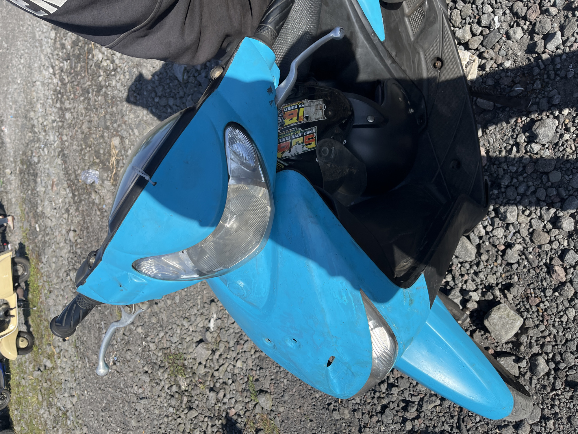 The image shows a blue scooter parked on a gravel lot. The scooter is in the foreground, with its handlebars and front fairing clearly visible. The front of the scooter has two circular holes and one elongated hole in the blue plastic. There are stickers on the black panel below the handlebars. In the background, a cream-colored scooter and a black scooter are partially visible, along with some trash on the ground. The environment appears to be an outdoor area with a lot of loose gravel.