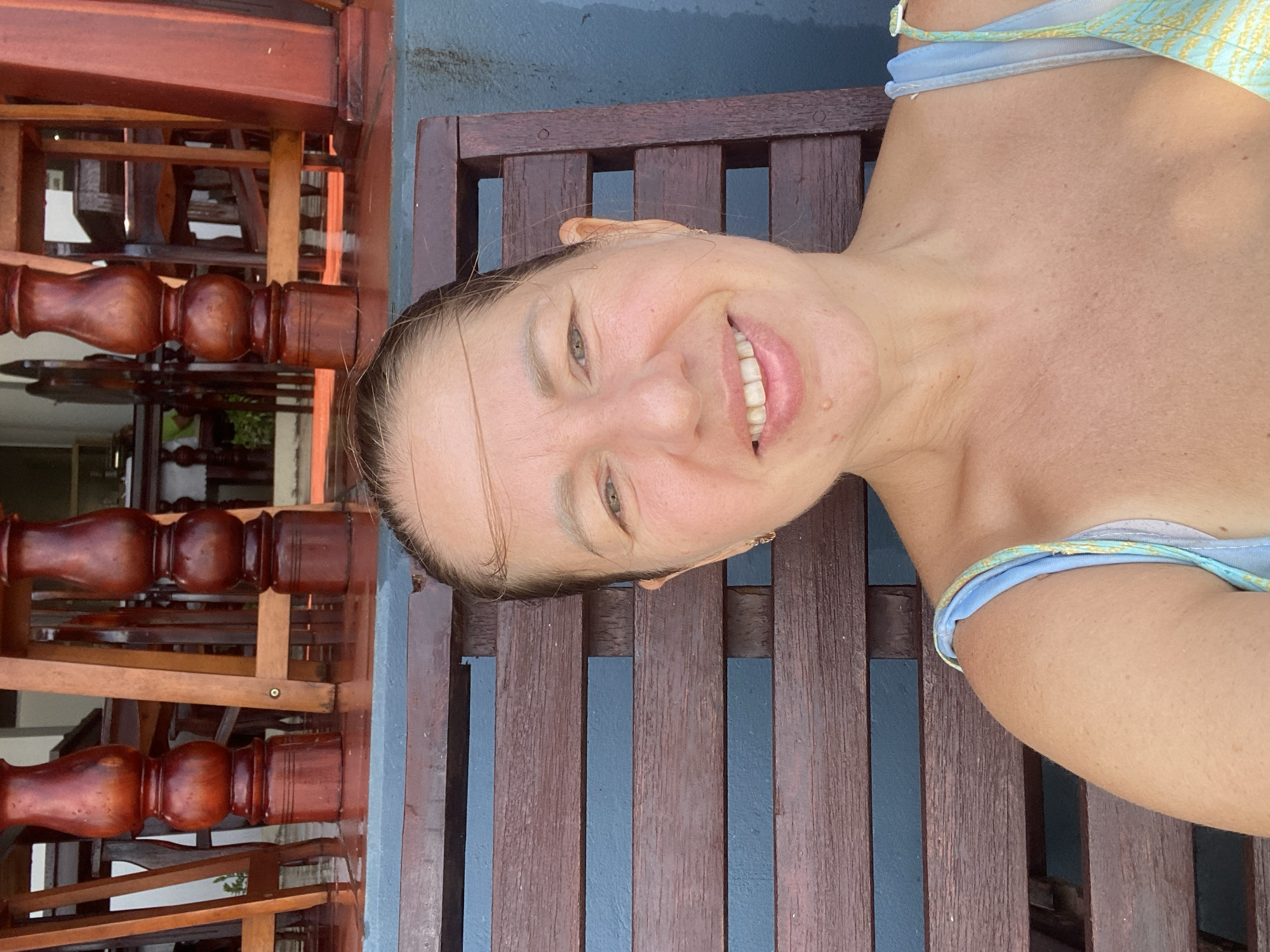 A woman is taking a selfie while sitting on a wooden bench. She has shoulder-length brown hair, a fair complexion, and is smiling. She is wearing a light blue and yellow floral print bikini top. The bench is made of dark brown wood with horizontal slats. Behind the bench, there is a blue wall and the legs of several wooden chairs and a table are visible, suggesting a restaurant or cafe setting. The floor in front of the bench appears wet and reflective, possibly from recent rain. The overall scene suggests an outdoor location in a warm climate, consistent with Mui Ne, Vietnam.