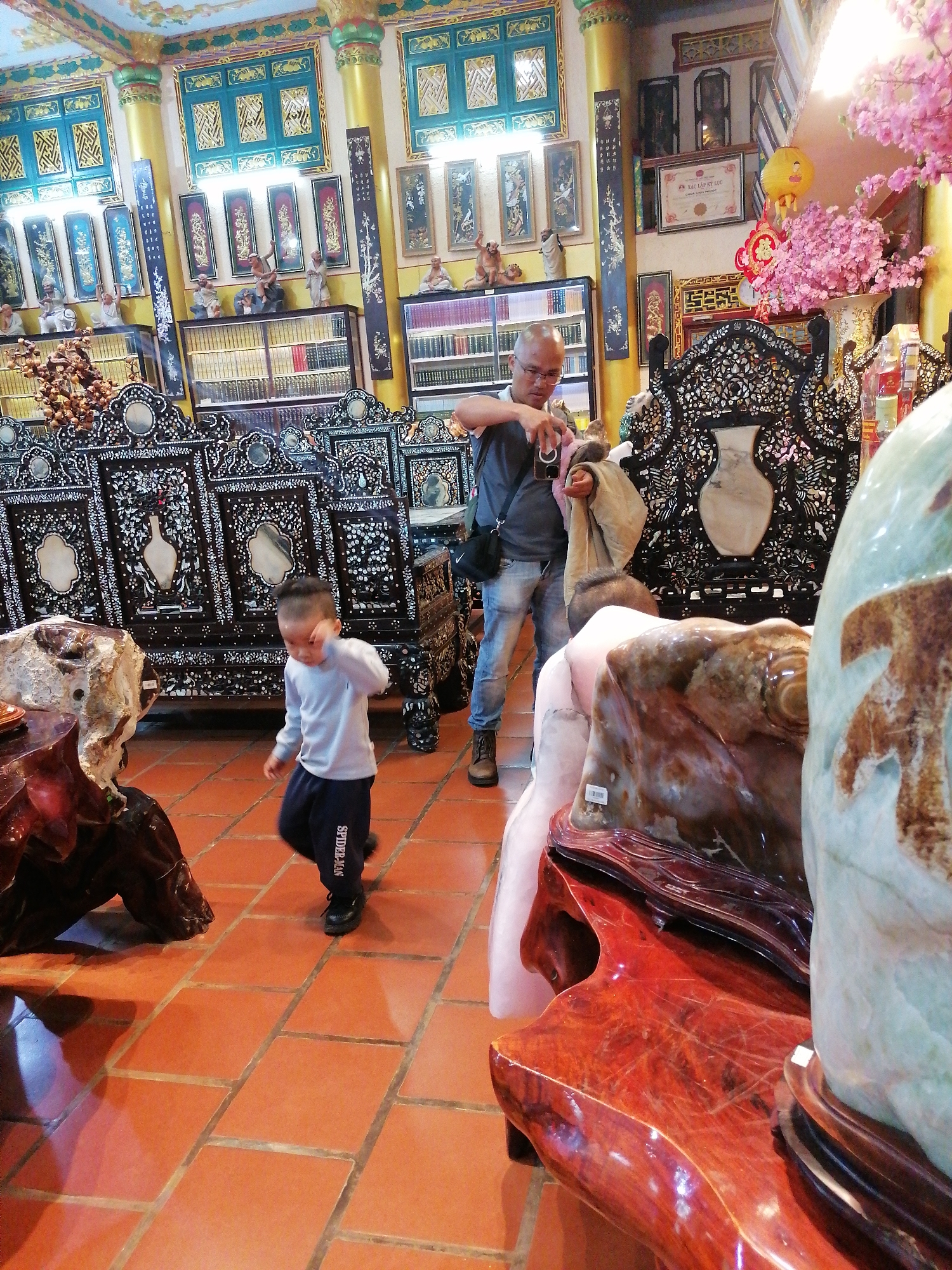 In Xuan Thanh city, Vietnam, a boy in a light blue long-sleeved shirt and dark pants with "SPIDER-MAN" written on the leg walks across a tiled floor. He is in motion, with his right arm raised and his head turned down and to the side. Behind him, a man in a grey t-shirt and blue jeans holds a camera up to take a photo. He is wearing glasses and has a bag slung over his shoulder. The environment is opulent, with ornate dark wood furniture inlaid with mother-of-pearl and intricate carvings. The walls are decorated with colorful paintings and framed calligraphy. A large, light blue jade-like sculpture is prominently displayed in the foreground, along with other large stone and wooden decorative pieces. Pink cherry blossoms are visible in the background. The overall impression is of a meticulously decorated shop or display space filled with cultural artifacts and decorative items.