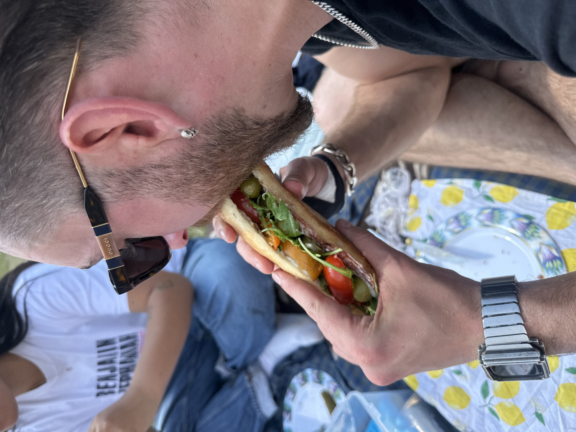 An image taken outdoors at ground level depicts a light-skinned person with dark hair and a beard eating a sandwich. The individual is wearing dark sunglasses with gold-colored frames, and a silver stud earring is visible in the ear. A silver-colored metal watch is on the person's wrist. The sandwich contains green olives, red tomatoes, green leaves, and possibly salami. Another person wearing a white shirt and blue jeans can be partially seen in the background. A patterned picnic blanket with lemons is spread out. The location is not specified and cannot be identified as London or the United Kingdom.