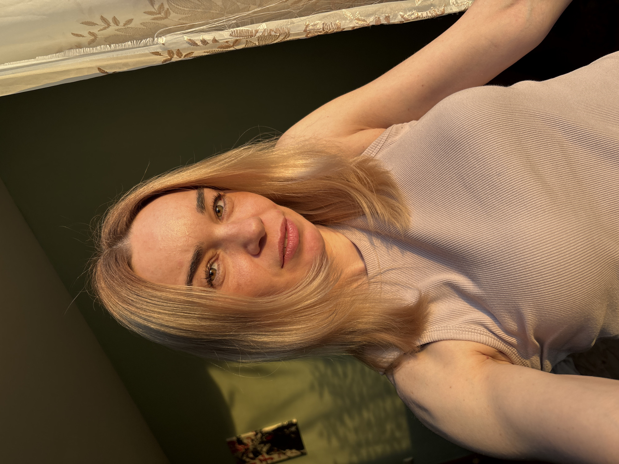 A woman with blonde hair and fair skin is depicted in a selfie-style shot, looking directly at the camera with a calm, neutral expression or slight smile. Her hair is parted and falls around her face and shoulders, highlighted by warm, golden light. She is wearing a light pink or beige ribbed tank top. The background behind her is a dark, muted green wall. Above her head, a sheer, light-colored curtain with embroidered leaf or floral patterns is visible, suggesting an indoor setting.
The lighting in the image appears to be natural sunlight, possibly from a window, casting warm highlights on her face and hair and creating soft shadows on the wall, indicative of late afternoon or early morning. Her eyes are light-colored, and her eyebrows are well-defined. Her lips have a natural pink hue. In the lower left background, partially obscured by shadow, a small, dark rectangular object with some lighter patterns, possibly a decorative item or a small picture frame, rests on a surface against the wall. There is no visible text in the image.