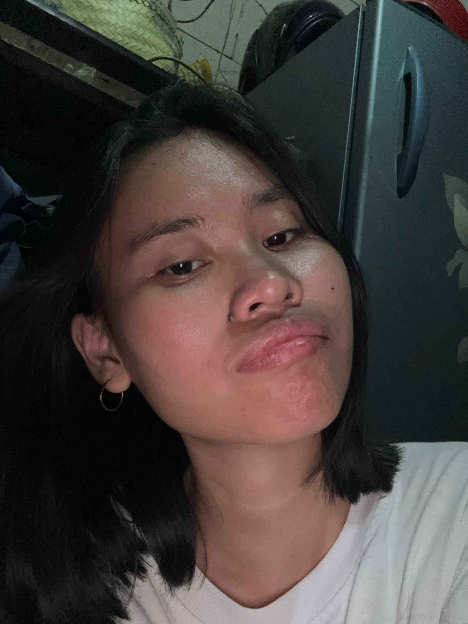 A close-up selfie shows a young woman with dark hair and a white t-shirt. She is looking directly at the camera with a slightly pursed expression. Her left ear, adorned with a small hoop earring, is visible, as is a nose piercing. The background appears to be an indoor setting, possibly a room, with some objects including what looks like a basket and a dark appliance, possibly a refrigerator. The lighting is dim, creating some shadows. The overall scene suggests a casual, personal moment captured indoors in East Jakarta, Indonesia.