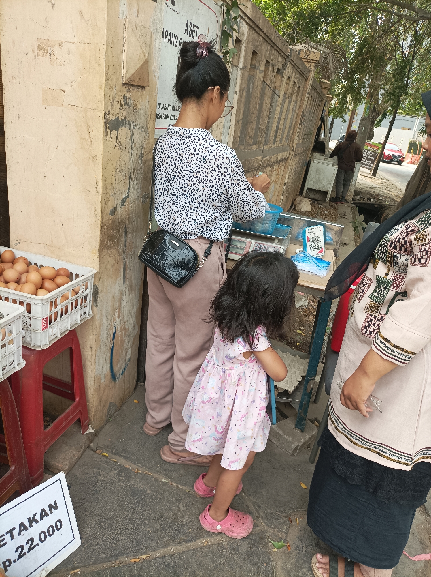 In a street scene in West Jakarta, Indonesia, a woman wearing a leopard-print shirt and light-colored pants stands at a makeshift stall. She has her hair tied up in a bun and is wearing glasses. Next to her, a young girl in a pink dress with a unicorn print watches something on the stall. A woman in a light-colored hijab and a patterned top stands to the right, observing the scene. The stall features a digital scale and a QR code for payment. Crates of eggs are visible on a red plastic stool to the left of the frame, and a sign with "ETAKAN Rp. 22.000" is on the ground in the foreground. The background includes a textured wall, trees, and a red car driving on the street.