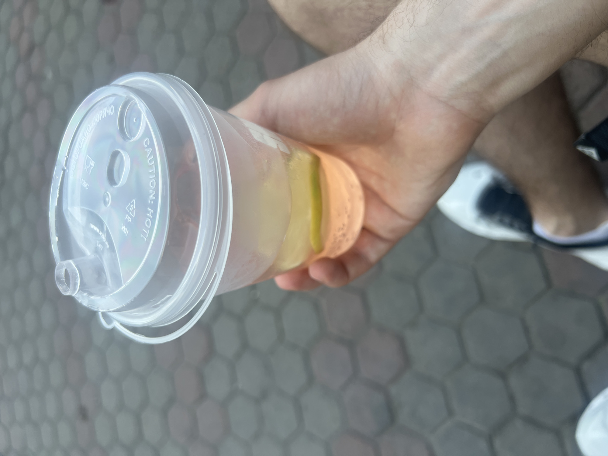 A person is holding a clear plastic cup with a lid, presumably containing a beverage, against a background of a grey hexagonal-patterned paving. The drink inside the cup appears to have layers of different colors, possibly indicating multiple ingredients. The lid has a protruding spout. The person's hand and part of their arm are visible, along with a glimpse of a shoe. The exact location in Krasnoyarsk, Russia cannot be determined from the image.