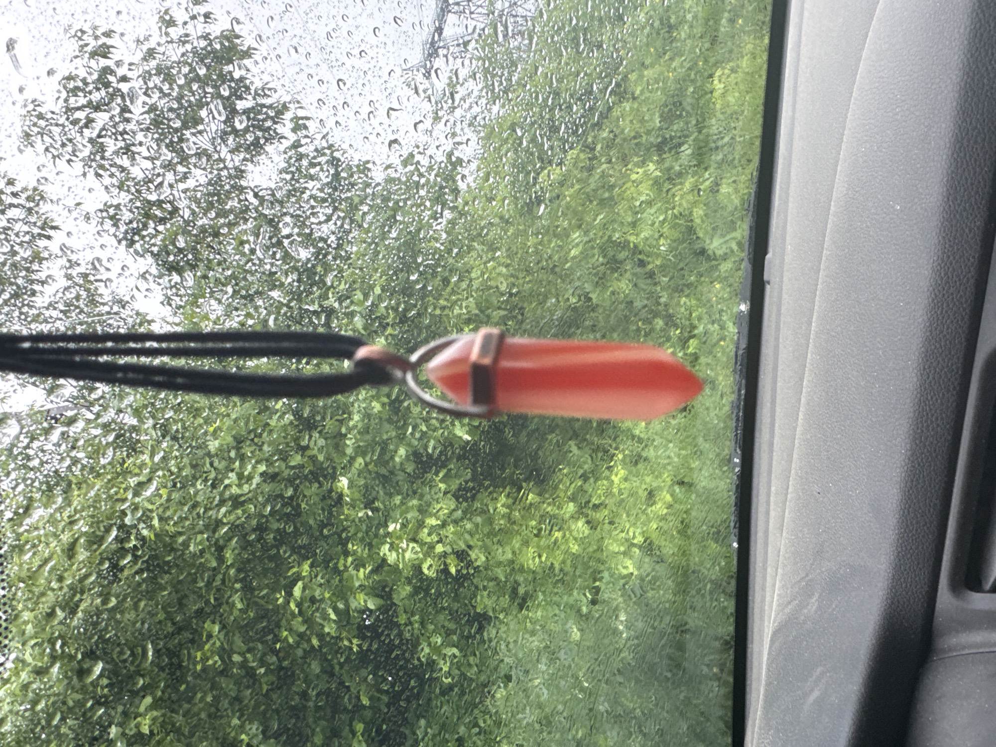 The image, taken through a rain-streaked window, features a red, pointed crystal pendant suspended from a black cord. The background consists of a blurred view of green foliage, possibly trees, and a grey interior car panel. The location of the image is unknown, and cannot be located based on the image alone. There are no people in the image.