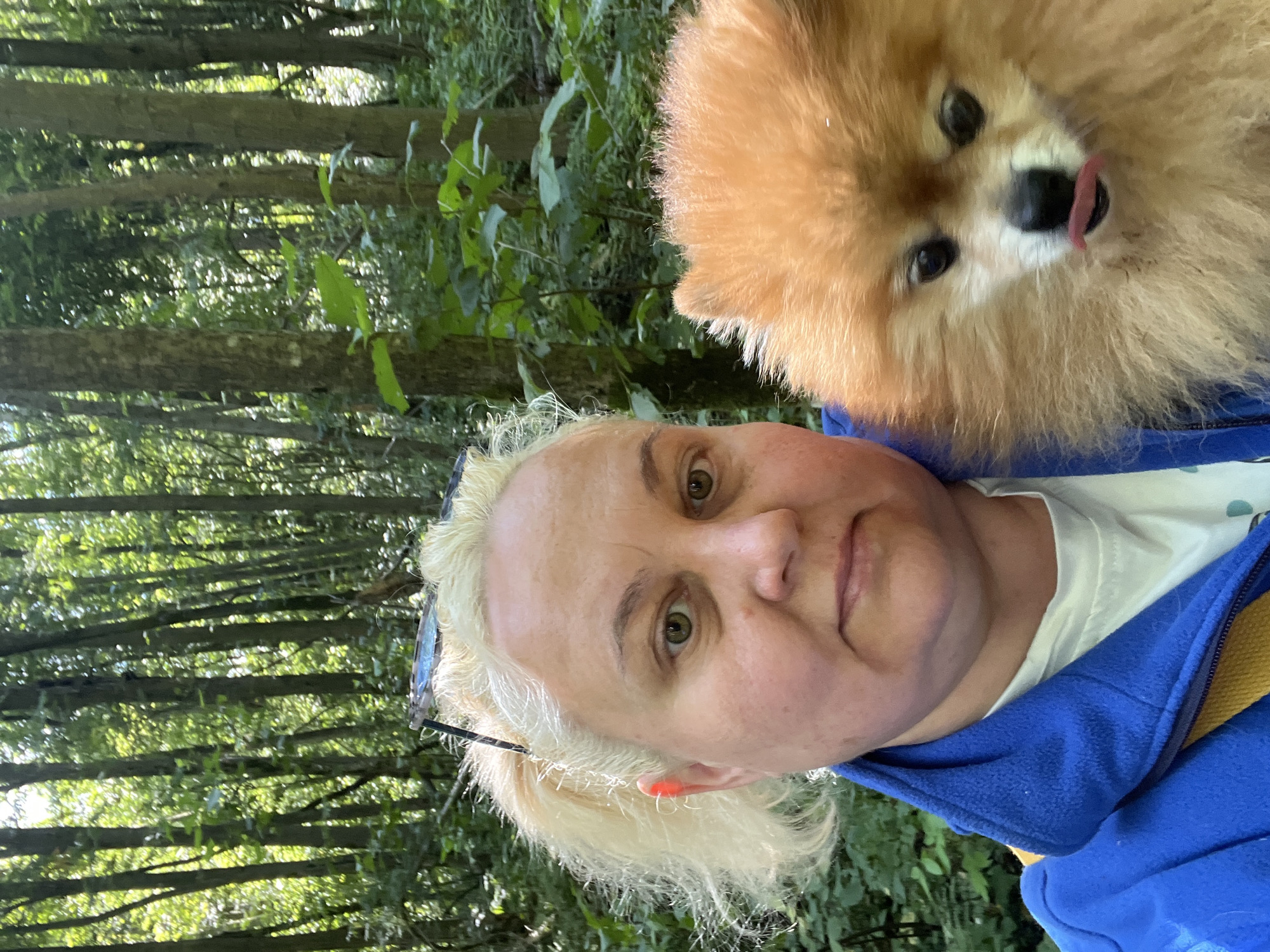 In a wooded area with a thick canopy of trees, a light-skinned woman with blonde hair pulled back in a ponytail takes a selfie. She is wearing a blue jacket over a white t-shirt. In her arms, she is holding a small, fluffy orange Pomeranian dog. The dog has its tongue sticking out and is looking at the camera. The background is out of focus, showing more trees and foliage. The overall lighting suggests it is daytime.