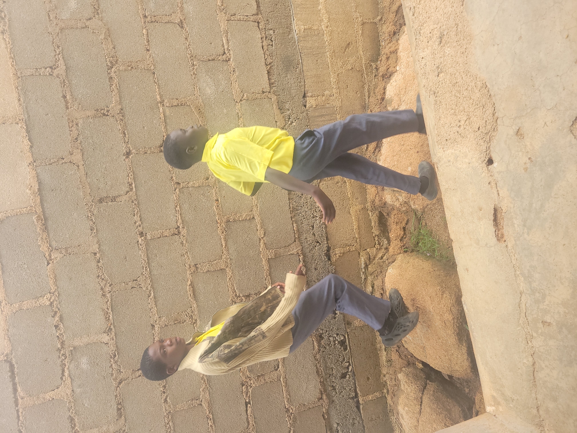 Two young males, possibly adolescents, stand in an outdoor environment. One wears a bright yellow shirt and dark trousers, while the other is dressed in a light-colored cardigan over what appears to be a yellow shirt, and similar dark trousers. They appear to be standing adjacent to a building constructed with concrete blocks. The background consists of similar structures and what could be ground cover. The image lacks specific markers to definitively place it within Jos, Nigeria, but the building materials and general environment are consistent with urban or peri-urban areas in Nigeria. The subjects are interacting, though the nature of their interaction is unclear from the image alone.