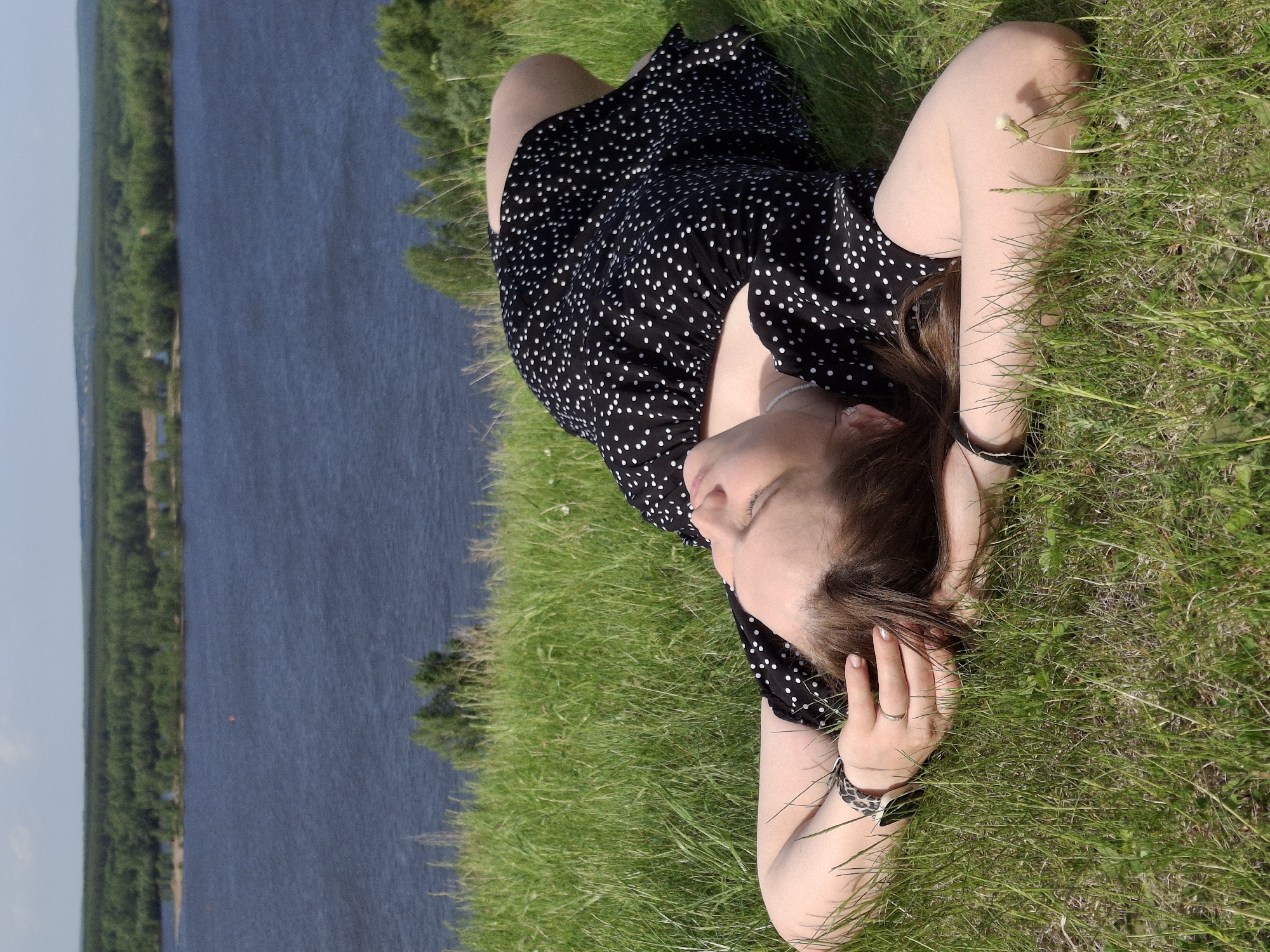 A woman is lying on a grassy hill overlooking a body of water in Ольховка, Russia. She is wearing a black dress with white polka dots. Her arms are positioned behind her head. The background features green trees lining the shoreline of the water.
