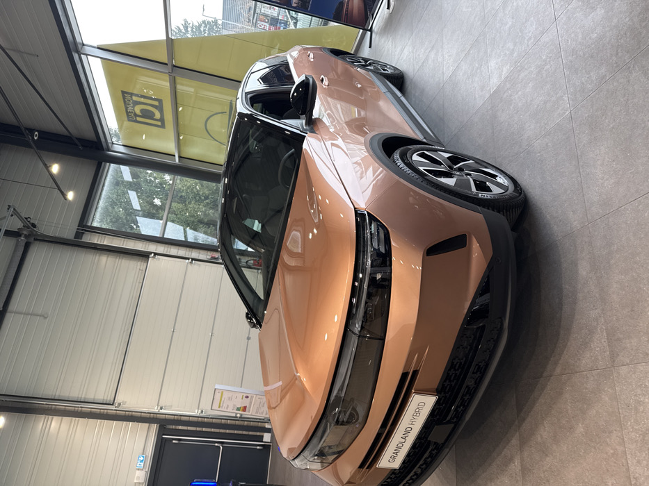 A metallic copper-rose gold Opel Grandland Hybrid SUV with a black roof and trim is displayed inside a car dealership showroom in Hoenheim, France. The vehicle, featuring a distinctive front fascia with integrated headlights and a "GRANDLAND HYBRID" badge on its lower grille, rests on a light gray tiled floor. The showroom environment includes a high ceiling with exposed metal beams and track lighting. To the left, a dark gray exit door and a large white sectional overhead door are visible, along with a yellow informational poster. To the right, large windows offer a view of trees and an external building. A prominent yellow sign, visible through or reflected in the windows, displays the mirrored French text "Nouveau ICI." No individuals are present within the scene, and the vehicle is stationary.