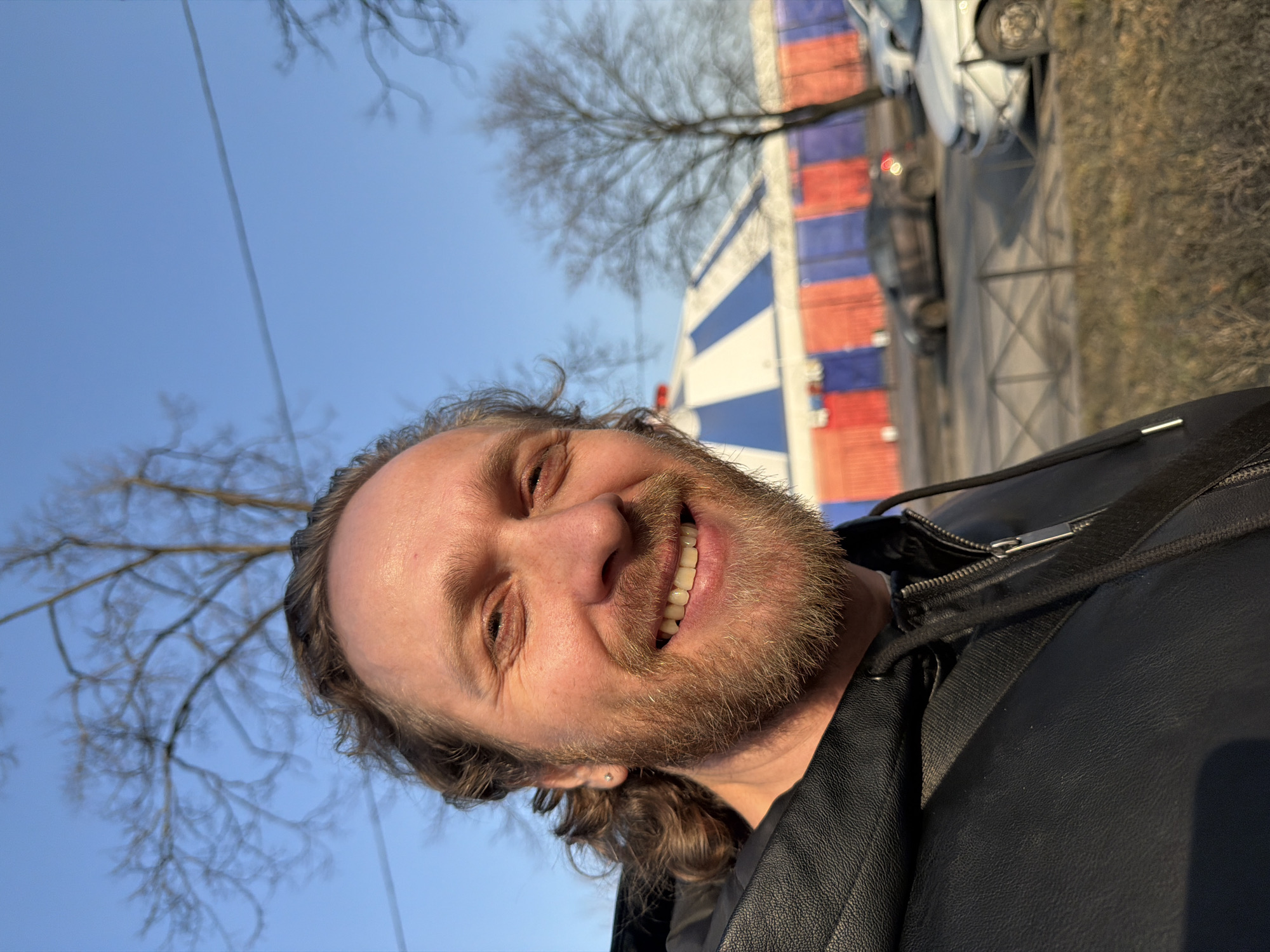 The image is a selfie of a man with fair skin, light brown hair, and a beard, smiling at the camera. He is wearing a black leather jacket with a visible zipper and strap. Behind him is a building with red, white, and blue horizontal stripes. In the background are trees without leaves, a blue sky, cars parked along a road, and a fenced area. The image was taken during the day. The location is likely Saint Petersburg, Russia, given the style of the building which is reminiscent of Russian architectural and color schemes.
