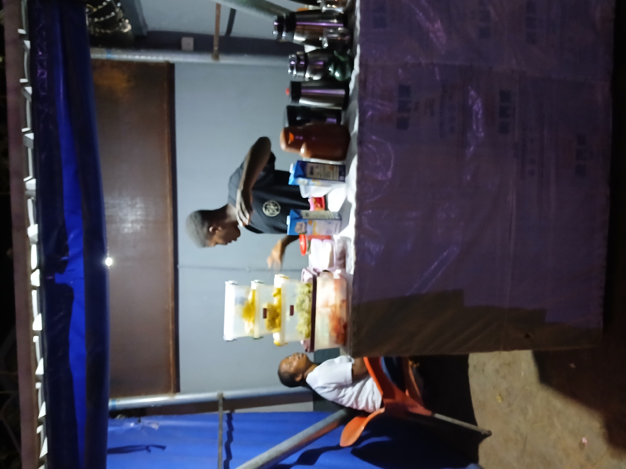 The image depicts an outdoor food stall in Bauchi, Nigeria. Two men are visible: one stands behind the counter wearing a black shirt, apparently preparing food or drinks. The other man is seated on an orange chair to the right, wearing a white shirt, possibly waiting or resting. The stall is covered with a light blue and purple tarp. In front of the counter are several clear containers filled with different food items. Various bottles and boxes are visible on the counter's surface, along with metal pitchers and appliances. The stall is constructed of metal poles. The surrounding area appears to be dimly lit, with minimal detail visible in the background.