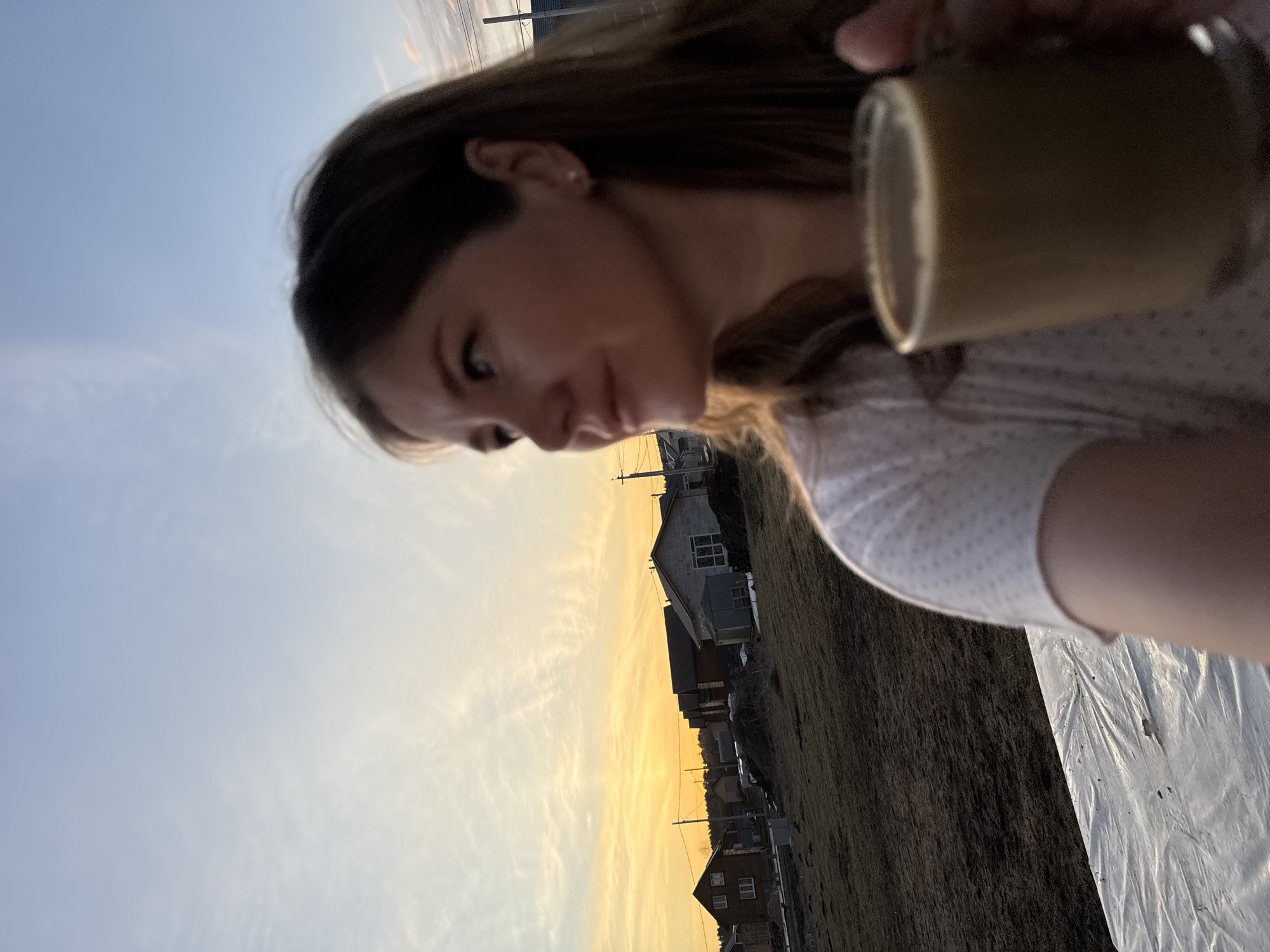 A woman with light brown hair, wearing a light-colored, polka-dotted top, is holding a brown mug. The background shows a sunset and a street of houses, possibly in Gornyi (Dvurechenskoe S/P), Russia, based on the presence of typical Russian residential buildings. There is also a plastic sheet in the lower right corner.