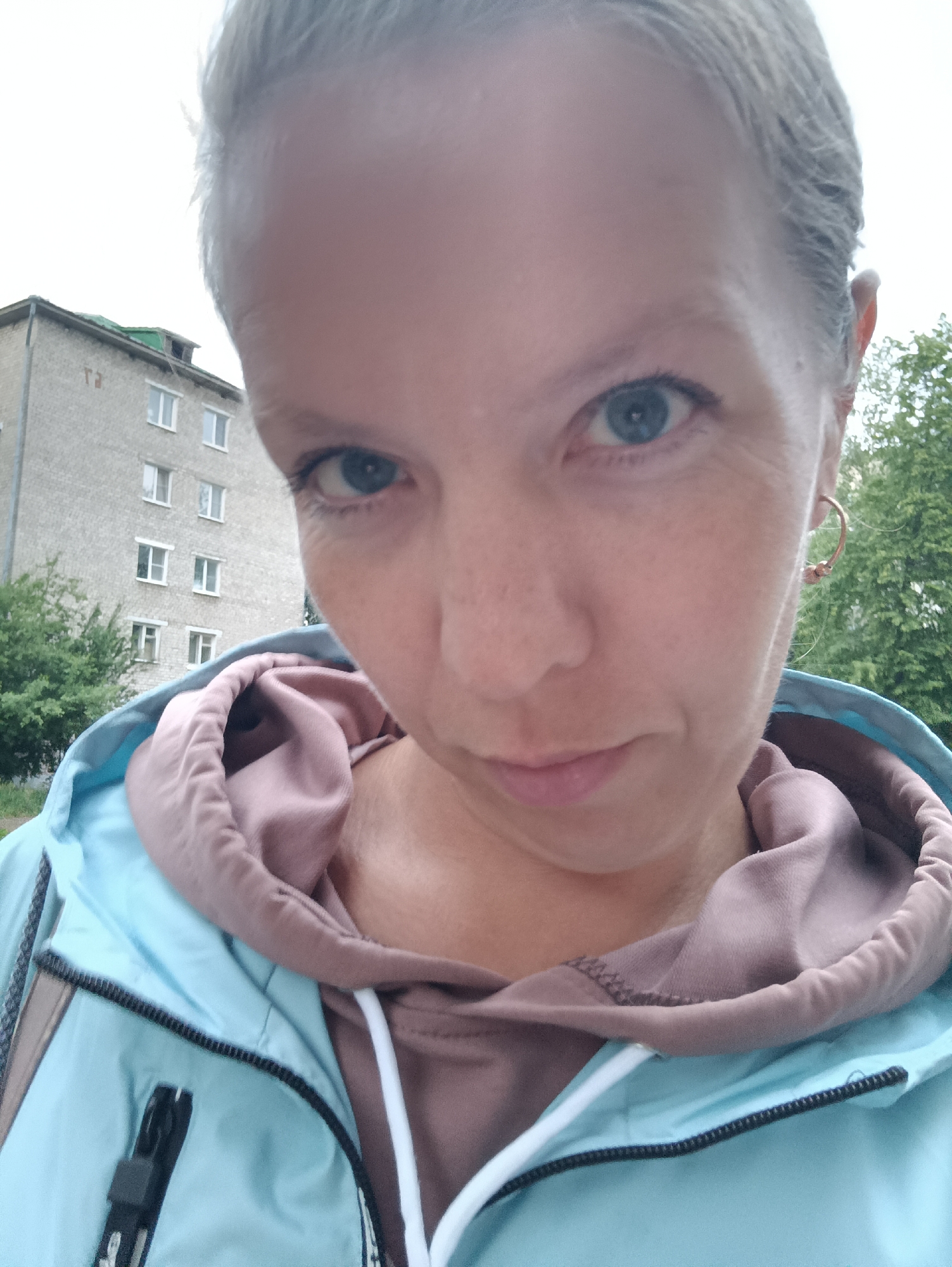 An adult female is depicted in a close-up, outdoor selfie taken in Chaikovsky, Russia. She has fair skin, short light-colored hair, and blue eyes. Fine lines are visible around her eyes, and subtle freckles are present on her face. She wears a light blue zip-up jacket, under which a light brown or dusty rose-colored hooded sweatshirt with white drawstrings is visible. A gold-toned hoop earring is present on her right ear. The background features a multi-story, light grey building constructed of concrete or brick, characterized by numerous white-framed rectangular windows. Faint characters, appearing as "тэ" or "TЭ" in Cyrillic, are visible on the upper left portion of the building. The building's roof includes a green section and a visible structural element. Green trees and bushes flank the building, indicating an outdoor setting under an overcast sky.