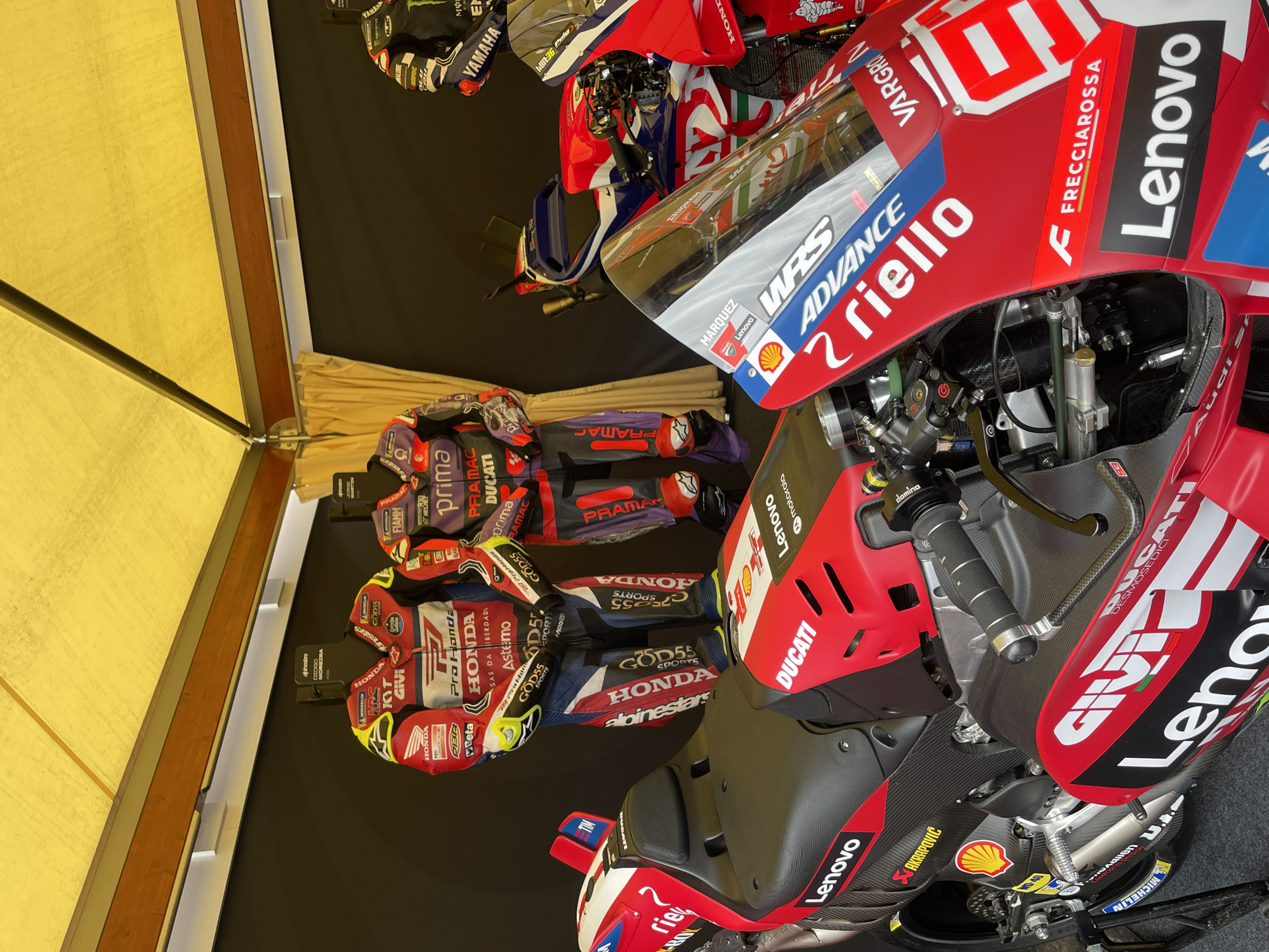The image depicts a display of motorcycle racing gear and bikes. The motorcycles are branded Ducati and Honda, with visible sponsorships from Lenovo, Shell, and Akrapovic. The racing suits and gloves displayed next to the bikes bear the same brands and sponsors, as well as Alpinestars and other logos. The setting is an indoor exhibit, likely a trade show or museum, with a yellow awning overhead. The image does not provide enough information to determine the location in Barcelona, Spain.