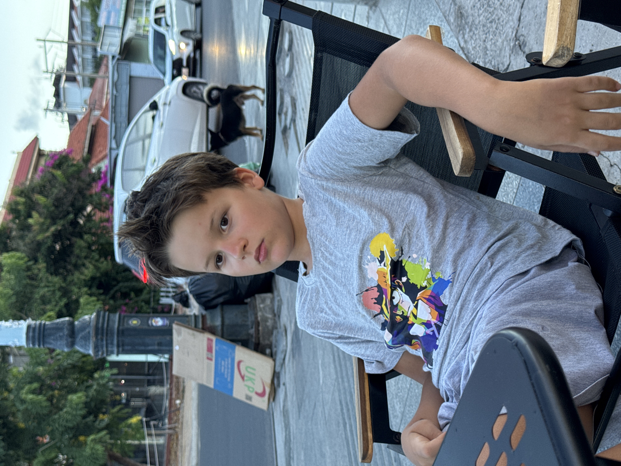 A young boy with brown hair and fair skin sits in a black chair with wooden armrests outdoors, wearing a gray t-shirt with colorful graphics. He is looking towards the camera. Behind him, a street scene in Phu Thuy Ward, Vietnam is visible. There are parked white cars, a black dog, trees, and streetlights, with buildings in the background. The overall lighting suggests it is daytime.
