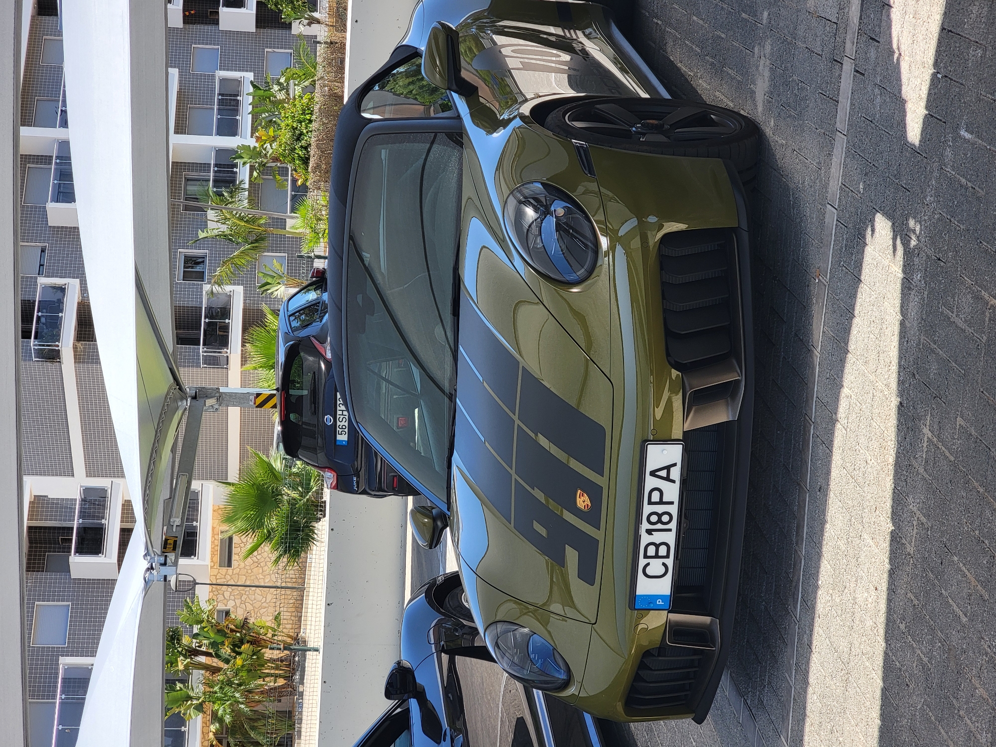 The image shows a close-up of a green Porsche 911 convertible parked in Quarteira, Portugal. The car has black racing stripes on the hood with the number "911" emblazoned on them. A Portuguese license plate with the characters "CB 18 PA" is visible. The car is parked on a paved surface, and a black Nissan Juke is visible in the background. A modern building with balconies and a tiled facade is situated behind the cars. Palm trees and other foliage are present in the surrounding area, suggesting a warm climate. The scene appears to be an outdoor parking lot, possibly associated with a residential or commercial complex in Quarteira.