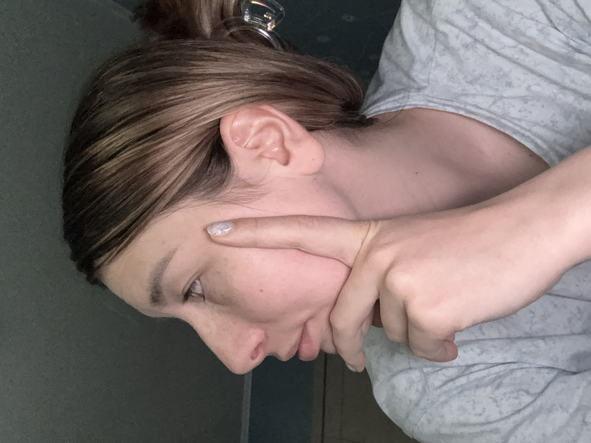A woman with light skin and brown hair pulled into a bun is shown from the side. She's wearing a light gray patterned shirt. Her right index finger is pointing towards her eye, and her other fingers are touching her cheek. The background is a solid, dark color. There is no evidence to suggest the location of this image is Moscow, Russia.