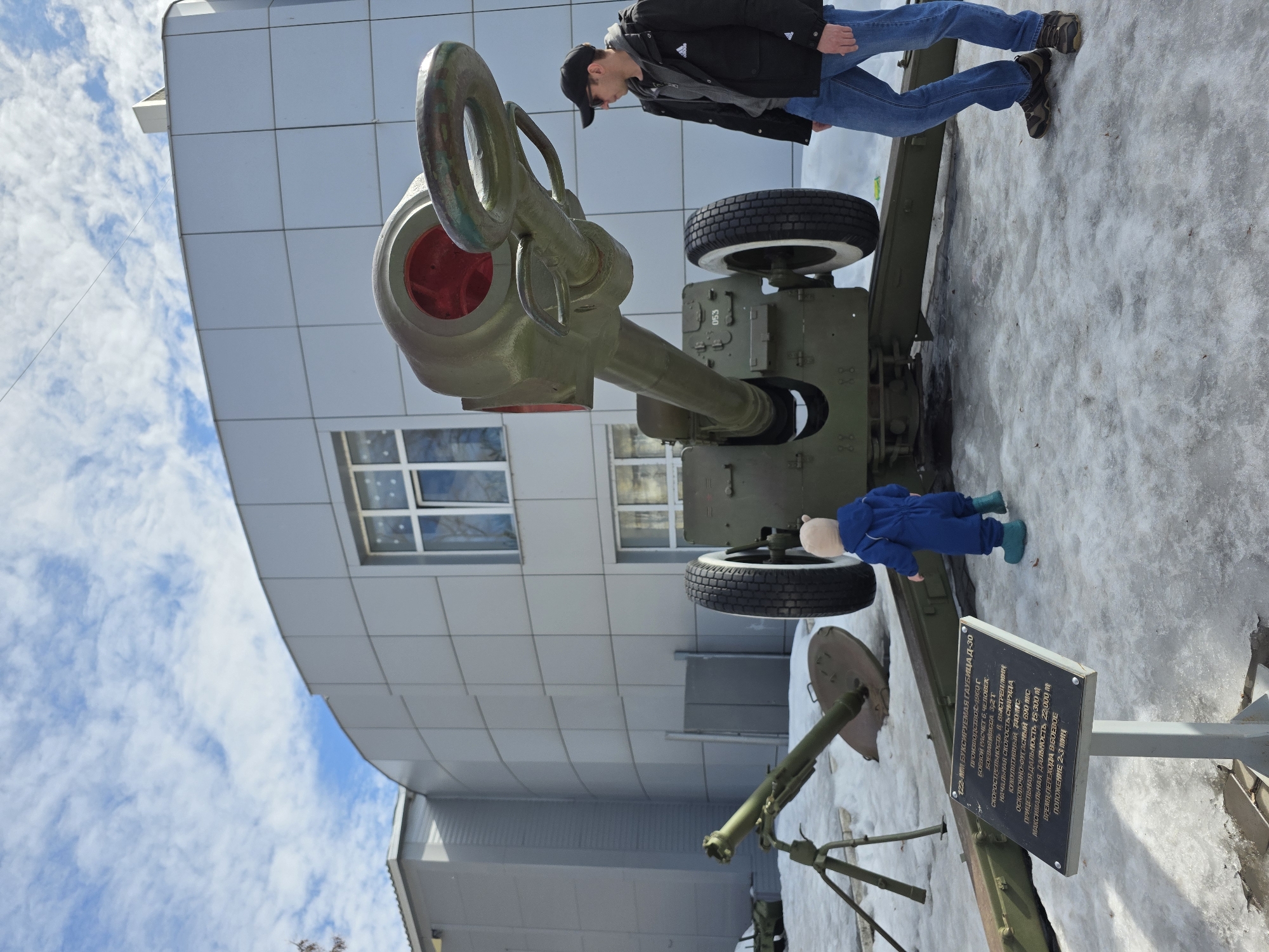 An outdoor scene in Votkinsk, Russia, depicts a display of military ordnance on a patchy snow and ice-covered ground. Dominating the center-right is a large, olive-green 122-mm D-30 towed howitzer, with its barrel angled upwards, revealing a red interior at the breech end. It rests on two large wheels with white sidewalls. To the left, a smaller olive-green mortar system, consisting of a barrel on a tripod and a circular base plate, is partially embedded in the snow. In front of the howitzer, a dark rectangular plaque mounted on a grey stand displays specifications of the "122-мм буксируемая гаубица Д-30" in white Russian text.
Two individuals are present: an adult male on the far right, standing beside the howitzer and facing left, wearing a black cap, sunglasses, a grey hooded sweatshirt, a black Adidas jacket, blue jeans, and brown hiking shoes. To his left, a young child, appearing to be a toddler, stands facing the howitzer, clad in a full blue snowsuit with a light tan hood and teal boots. Neither individual is actively interacting with the equipment beyond observation.
The background features a modern building with a light grey paneled exterior, a prominent curved section, and visible windows. The sky overhead is blue with scattered white cumulus clouds.