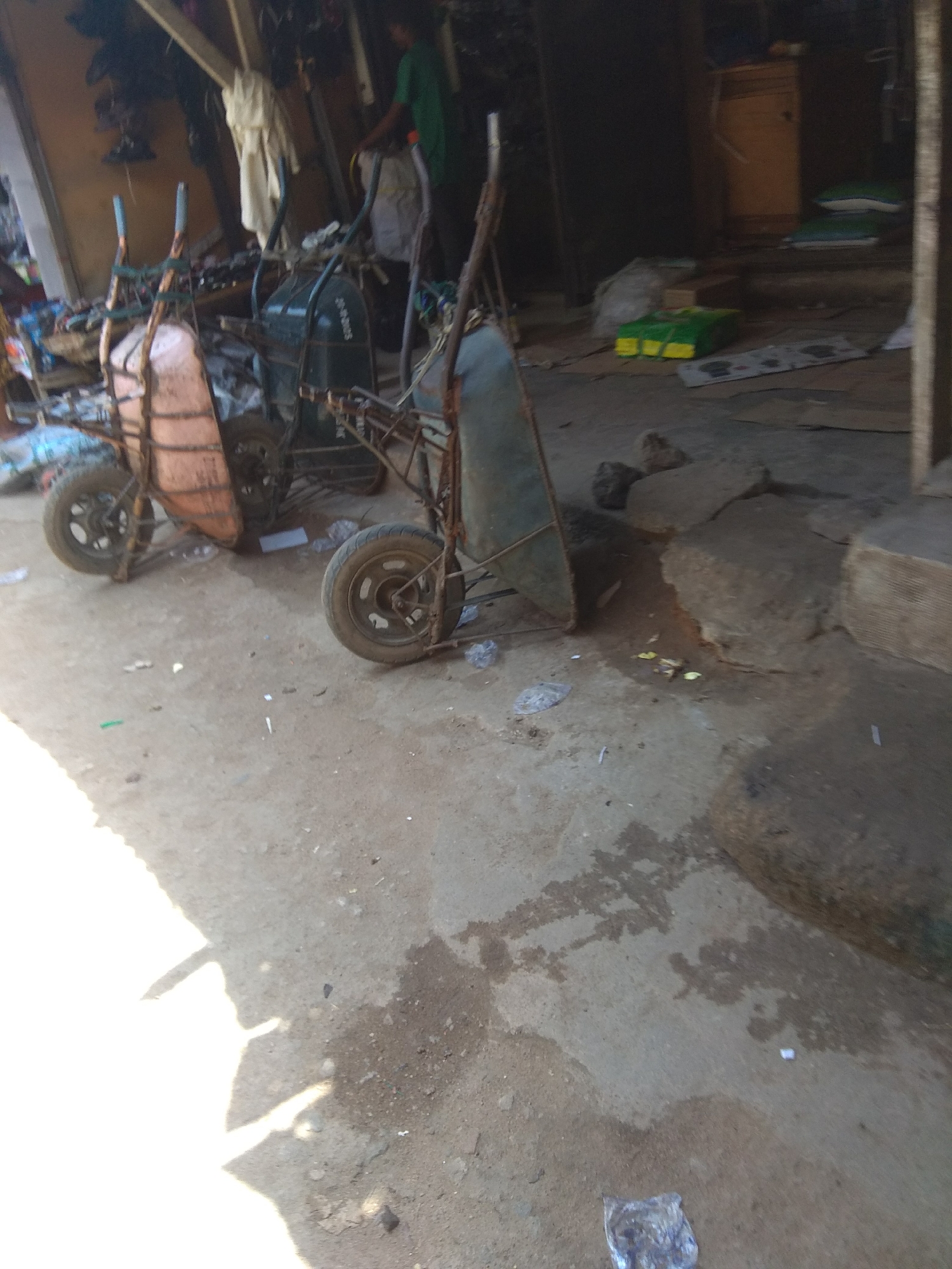 The image depicts an outdoor scene in Jalingo, Nigeria, featuring two wheelbarrows parked on a dirt and concrete ground. The wheelbarrows appear to be old and somewhat rusted. One wheelbarrow has an orange-colored bucket, while the other has a green bucket. In the background, a person wearing a green shirt can be seen in what appears to be a workshop or a storage area. The setting is cluttered with various items, including what looks like sacks or bags, fabric, and debris scattered on the ground, suggesting a busy or perhaps unkempt environment.
The lighting suggests it is daytime, with a strong light source casting shadows on the ground, particularly on the left side of the image. The overall atmosphere is utilitarian and appears to be a typical day in a working area. There is no specific event clearly taking place, but the presence of the wheelbarrows and the person in the background implies ongoing work or preparations for work. Some scattered litter is visible on the ground, adding to the unpolished feel of the location. There is no visible text in the image that provides specific information.