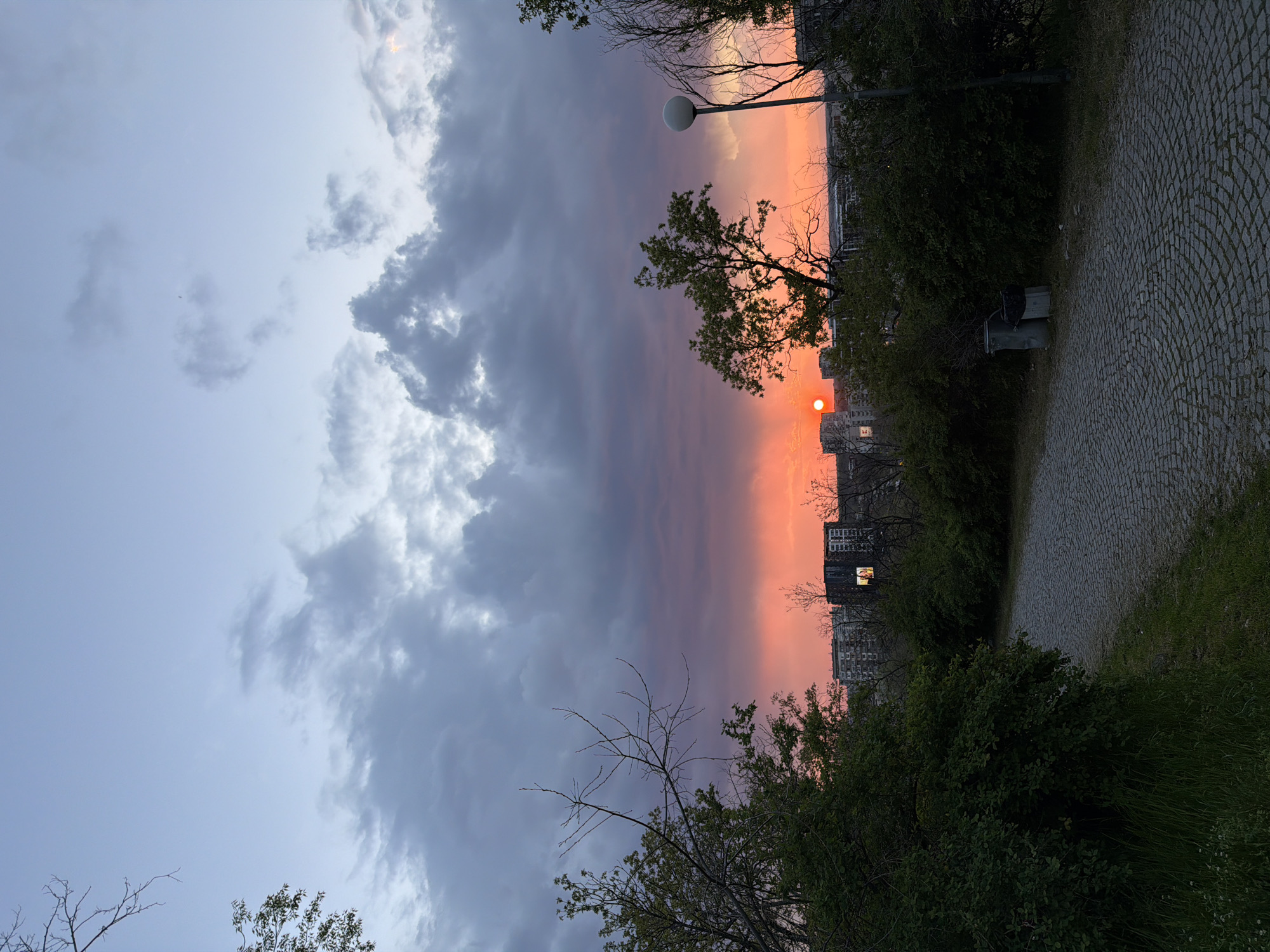 The image captures a sunset scene with a distinct division in the sky. To the left, the sky is pale blue with grey clouds, while the right side is dominated by orange and red hues of the setting sun. The sun is visible as a red orb on the horizon.
The foreground consists of a tree line with some buildings silhouetted against the sunset. A lamppost is also visible. To the right, a path made of grey stones.
Given the presence of buildings and greenery, this scene might be a park or a recreational area within the city of Plovdiv, Bulgaria.