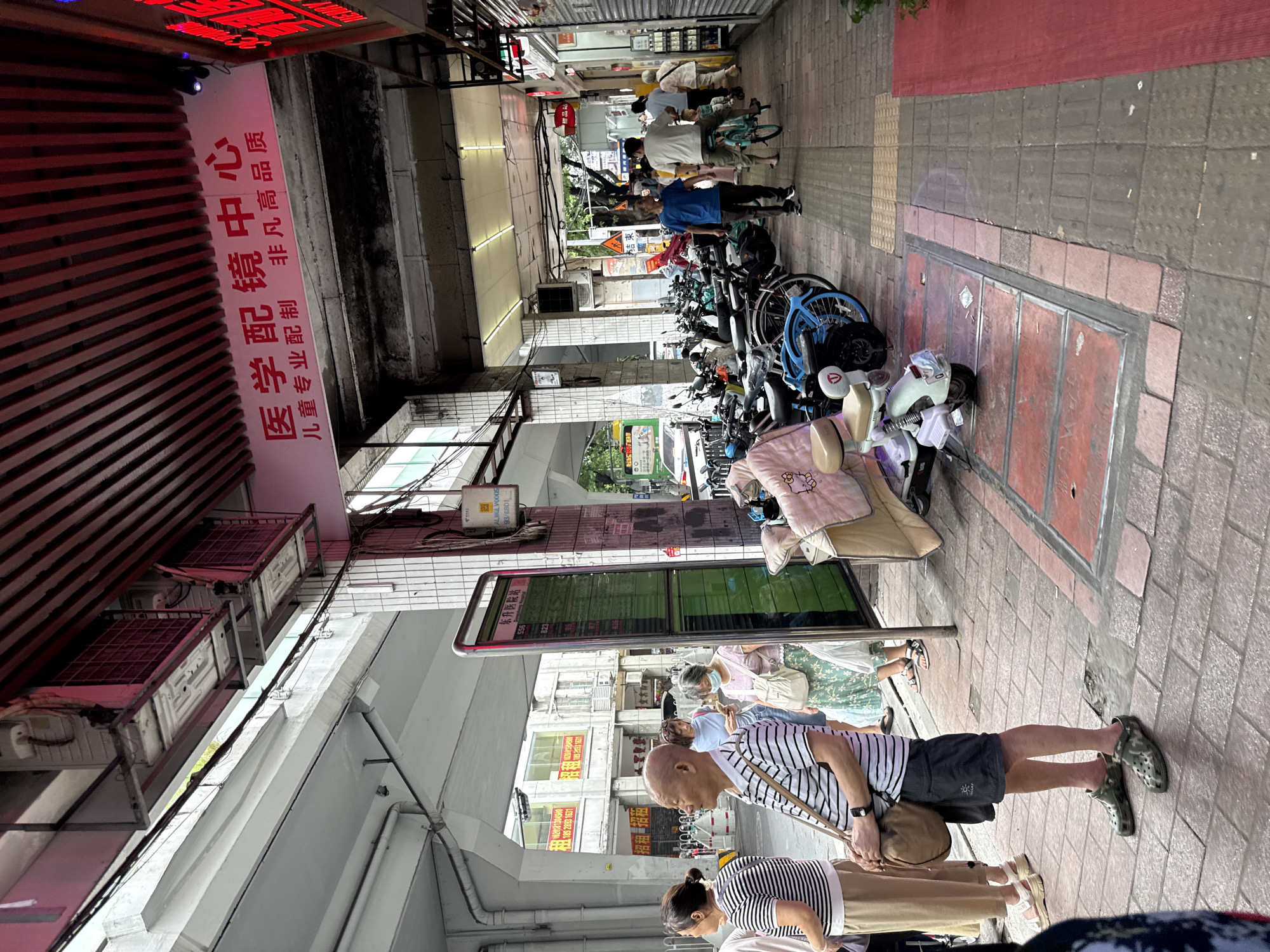 In Yuexiu District, China, a busy sidewalk scene unfolds under an overhead structure. A tall, digital display board, likely for public transport, stands prominently on the right side of the walkway, showing a green interface with rows of text and numbers. Adjacent to it, a column displays various signs, including what appears to be a building or establishment name in red lettering at the top, and a white sign with Chinese characters and a barcode at the bottom.
Several people are present, some waiting near the display board. A man in a striped shirt and dark shorts stands in the foreground, his gaze directed forward. To his left, a woman in a striped shirt and light-colored pants waits patiently. Further back, a woman in a blue top and a woman in a floral skirt and pink top are visible. A man in a blue t-shirt walks along the sidewalk, and another man in a white t-shirt rides a bicycle, a young child behind him. In the distance, more individuals and a bus are partially visible.
The sidewalk is paved with interlocking bricks. To the left of the display board, a covered area with pillars supports the overhead structure, which is lined with horizontal red slats. The ground in front of the covered area is also paved with bricks, with some metal grates or covers embedded in it. Numerous parked motorcycles and bicycles line the walkway, with some displaying blankets or covers. On the right side of the walkway, a red carpet or mat runs along the edge of a building facade which has several doors and windows, and displays various signs and merchandise. The overall environment suggests a commercial street with a focus on transportation and pedestrian activity.