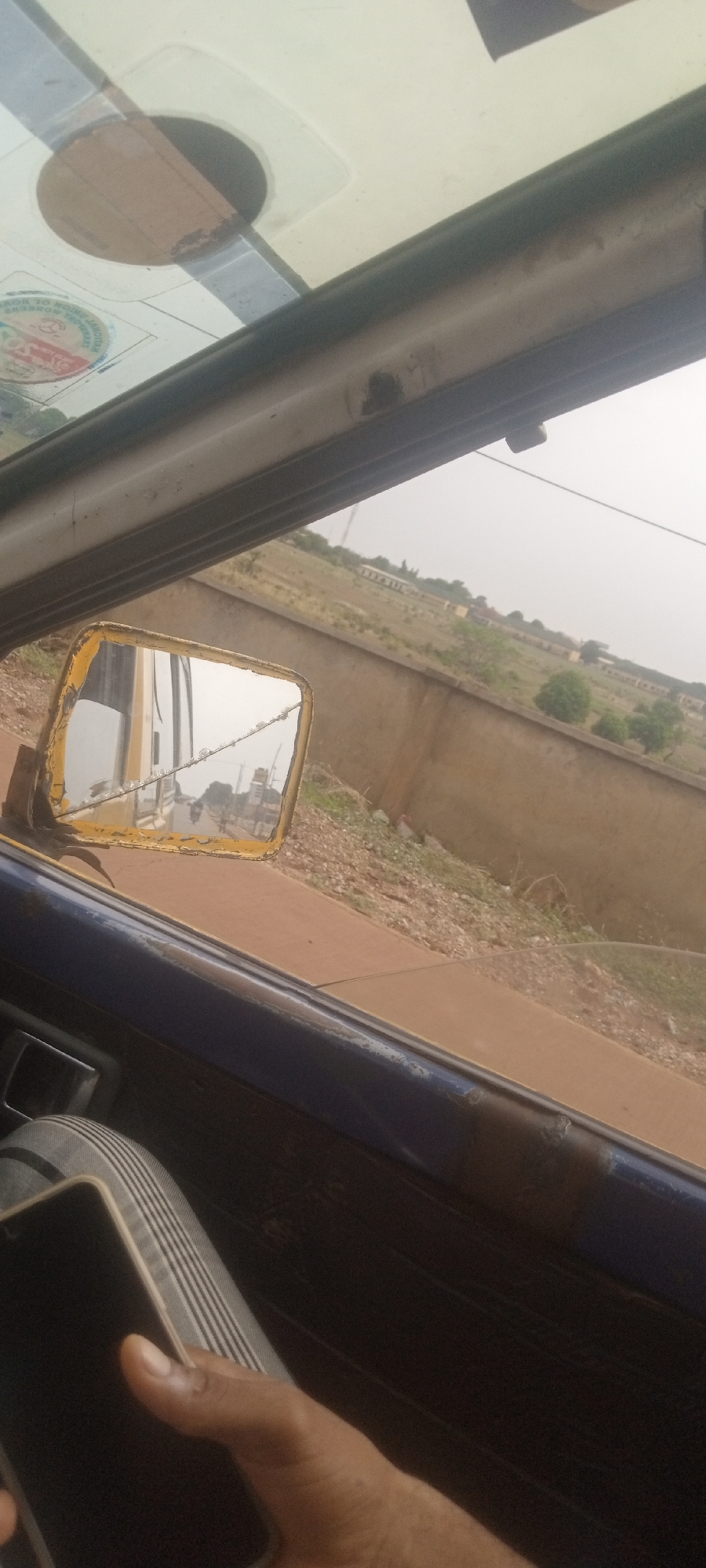 The image depicts an interior view from a moving vehicle, likely a taxi or bus, with the focus on the outside scenery and a cracked side mirror. The mirror reflects a street with a passing motorbike. The exterior shows a raised, weathered concrete wall bordering a grassy field, possibly on the outskirts of the city. The sky appears overcast and hazy. A hand holding a mobile phone is partially visible in the lower left corner of the image. The vehicle's interior shows signs of wear and tear. The setting appears to be in a rural or developing area. No specific location in Television, Nigeria is discernible, but the scene suggests a location on the outskirts of the city.