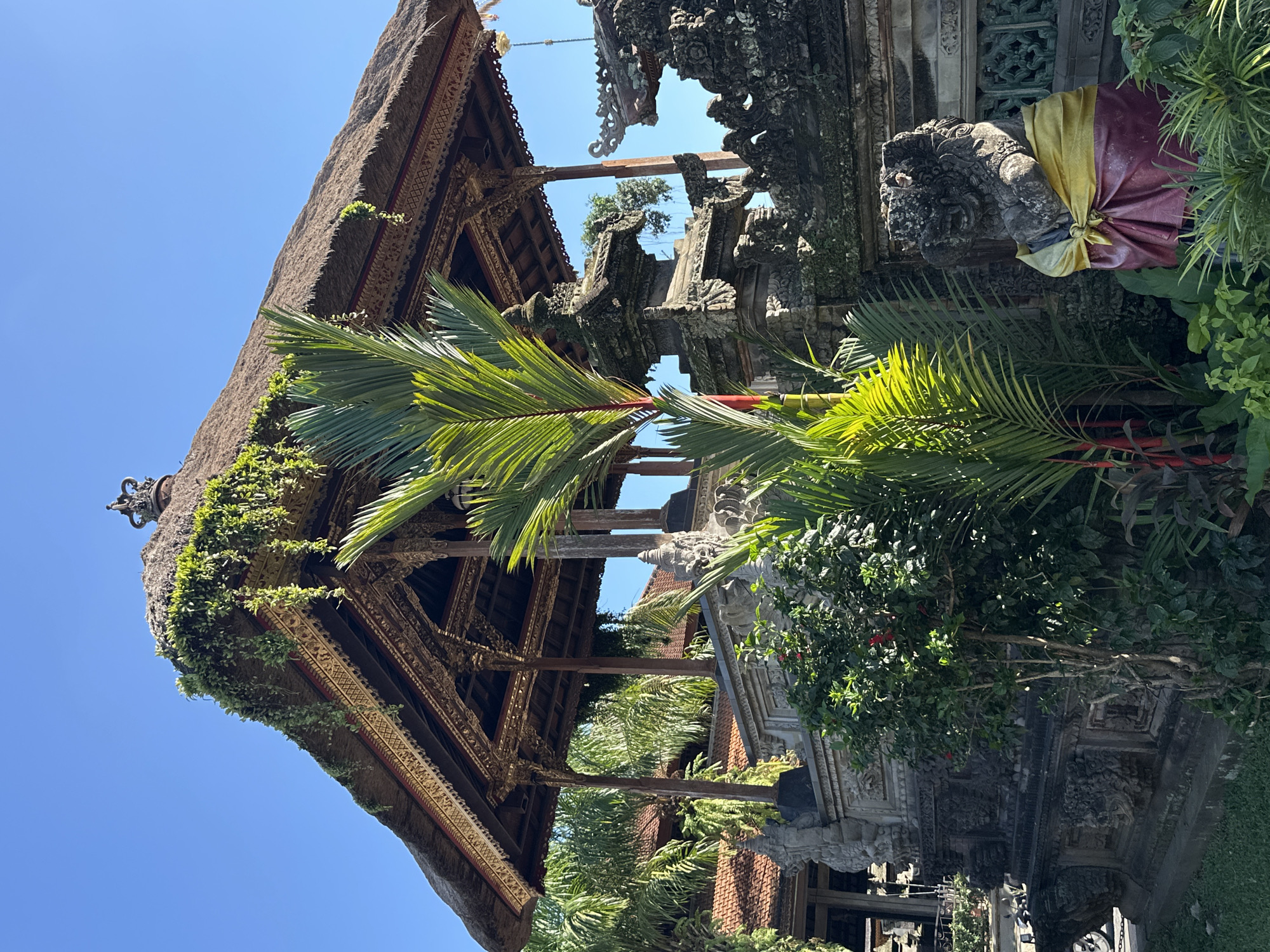 A statue of a lion-like creature, adorned with a yellow and red sarong, stands guard in front of an elaborately carved stone wall. Behind the wall, a gazebo with a thatched roof, overgrown with vines, is supported by intricately carved wooden pillars. Lush green foliage, including palm fronds and flowering plants, surrounds the structures, creating a vibrant tropical scene under a clear blue sky. The architectural style suggests a location in Ubud, Indonesia, likely a temple or traditional Balinese compound.