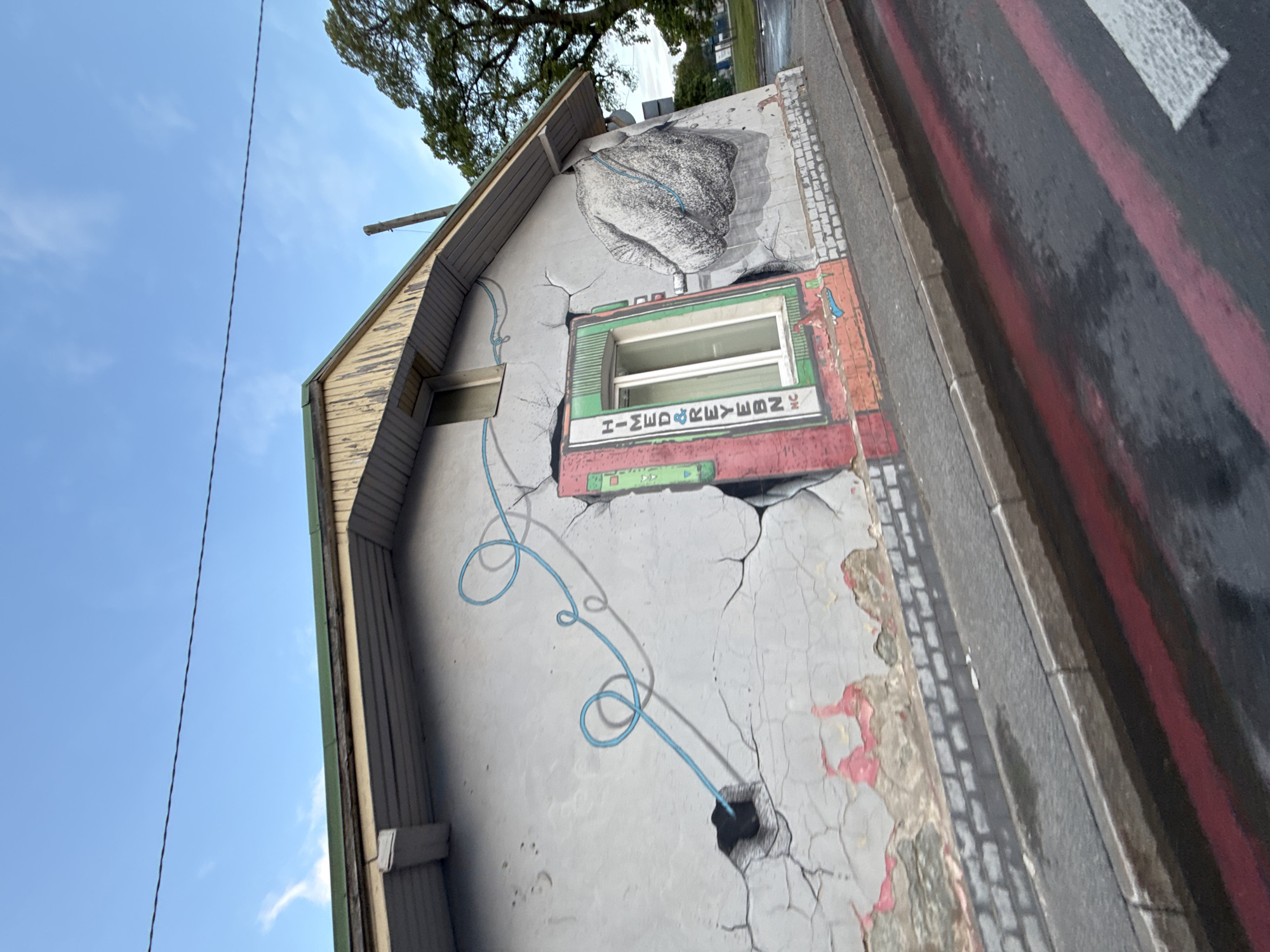 The image shows a street view in Tallinn, Estonia. The focal point is the side of a building with an artistic mural painted on its grey, cracked plaster wall. The mural depicts a large, textured grey hand reaching towards the right of the frame, and a blue, coiled line that appears to be connecting to a hole in the wall on the left. A green-framed window is incorporated into the mural, with the text "HIMED&REYBN, NC" vertically printed on it. The building has a yellow, peeling wooden soffit and a green roof edge. Below the wall, a sidewalk with a brick pattern is visible, transitioning to a wet asphalt road with red markings. In the background, trees and a clear blue sky with sparse clouds are present. There are no people visible in the image.