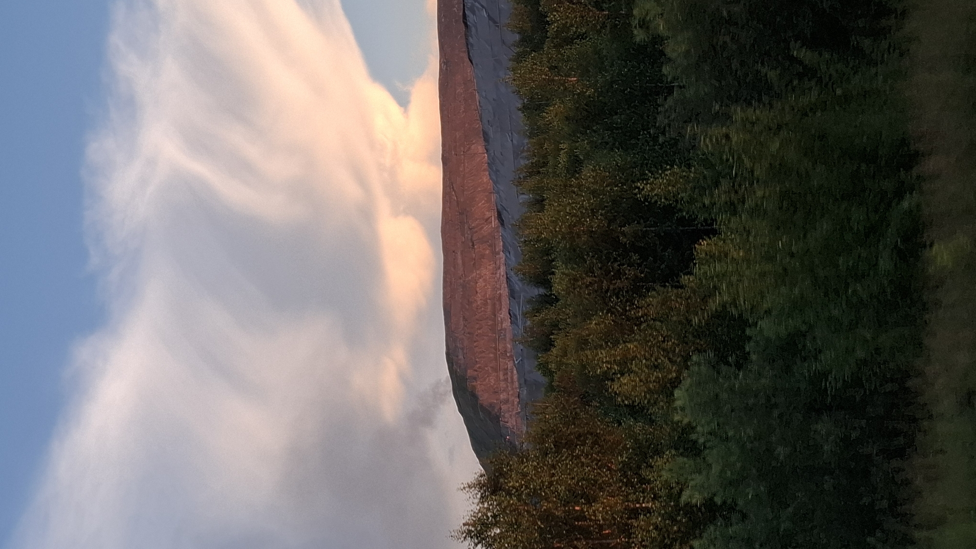 The image shows a vertical rock face with a forest in the foreground. The rock face is illuminated by the setting sun, casting a warm, reddish glow. The sky above is a soft blue with wispy clouds tinged pink and orange. The forest is lush and green, with trees of varying heights. The image appears to have been taken during golden hour, as indicated by the warm lighting and the colors in the sky. The location is Achinsk, Russia, though there are no specific landmarks visible to confirm this. The overall mood of the image is serene and picturesque, evoking a sense of natural beauty and tranquility. No people or text are visible in the image.