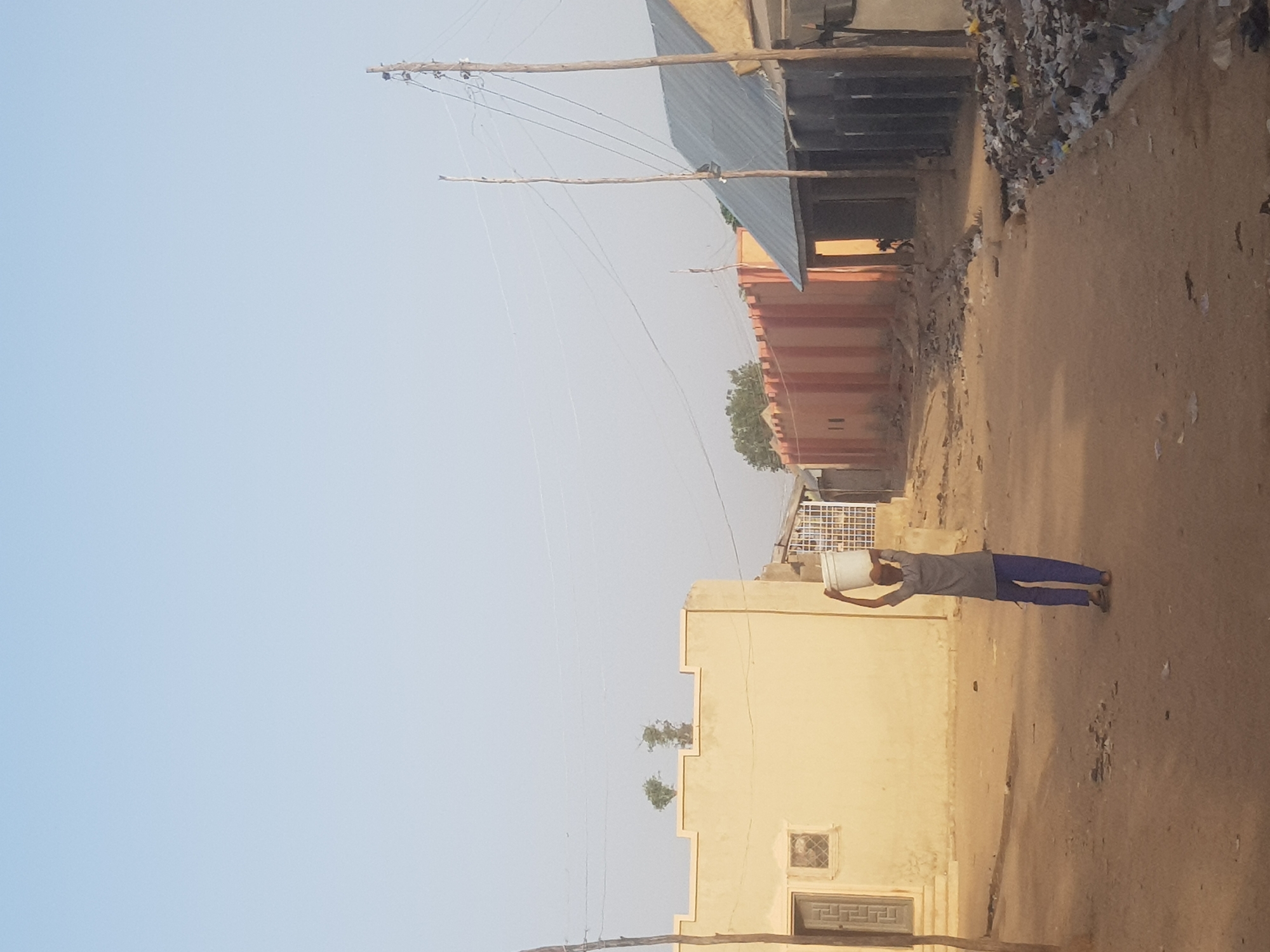 A man balances a white bucket on his head while walking down a dirt street in Dutsinma B, Nigeria. The street is lined with buildings. Power lines extend overhead.