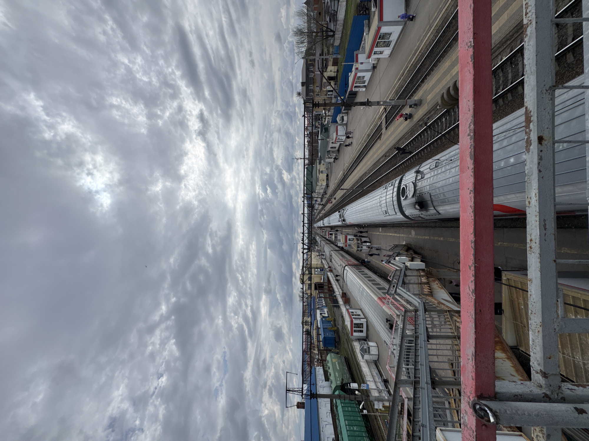 This photograph, taken in Rossosh, Russia, provides an elevated view of a railway station under a cloudy sky. Multiple railway tracks run parallel through the scene, occupied by parked passenger trains. On one side of the tracks, there are buildings, including small, prefabricated structures, and possibly residential or commercial buildings in the distance. The platform appears to be populated with several people.
In the foreground, part of a pedestrian overpass or bridge is visible, constructed from steel with sections painted red and gray. The perspective suggests the photo was taken from this structure. The cloud cover dominates the upper portion of the image.