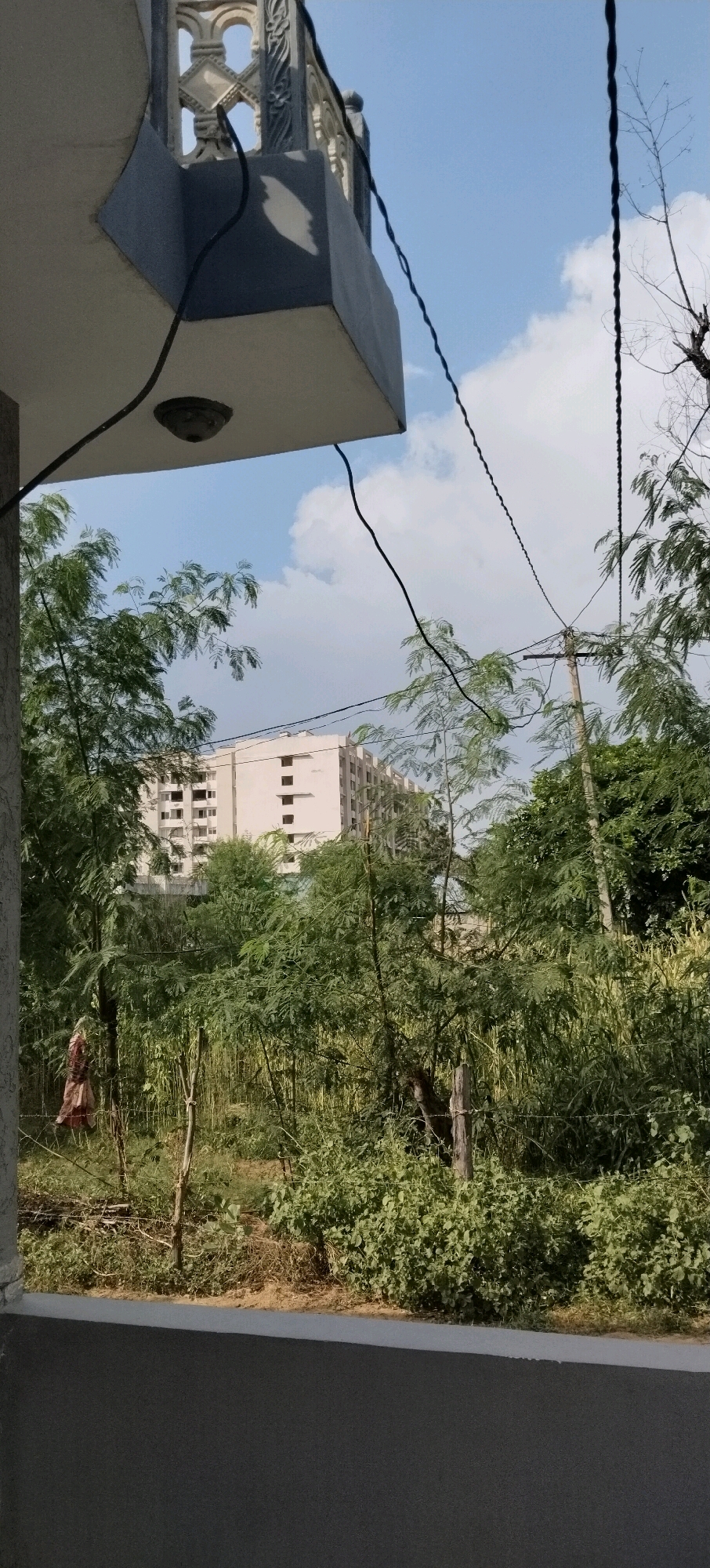 The image captures an outdoor scene on a sunny day in Mahapura, India. In the foreground, a gray concrete balcony railing is visible, with a black security camera mounted on the underside of an overhang. Wires traverse the scene, connecting to utility poles. Beyond the balcony, a dense green foliage of trees and bushes dominates the midground. In the distance, a large, light-colored building with multiple floors and rows of windows stands against a blue sky with scattered white clouds. A figure, appearing to be a person wearing a reddish-brown garment, is partially visible on the left side of the frame, standing amidst the greenery. The overall impression is of a residential area with ongoing construction and natural elements.
