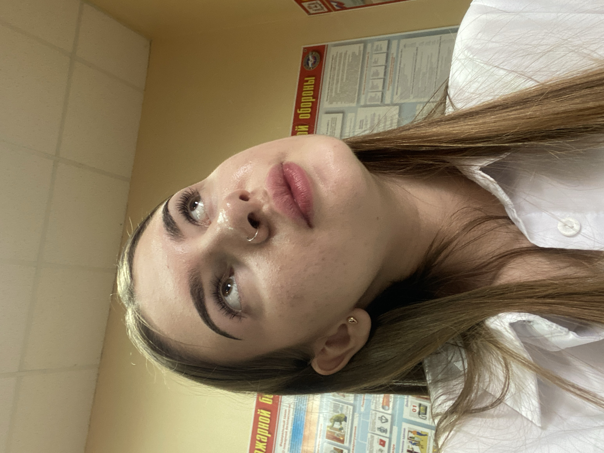 A young woman with light brown hair and light skin looks upwards. She has thick eyebrows, noticeable eyelashes, and a small hoop nose ring. Her lips are slightly parted and painted pink. She is wearing a white collared shirt. In the background, a yellow wall is visible, with posters mounted on it. One poster has red text that reads "ПОЖАРНОЙ БЕЗОПАСНОСТИ" which translates to "FIRE SAFETY". Another poster has red text that reads "ОЙ ОБОРОНЫ" which translates to "DEFENSE". The location is stated to be the city of Дьяково, Russia.
