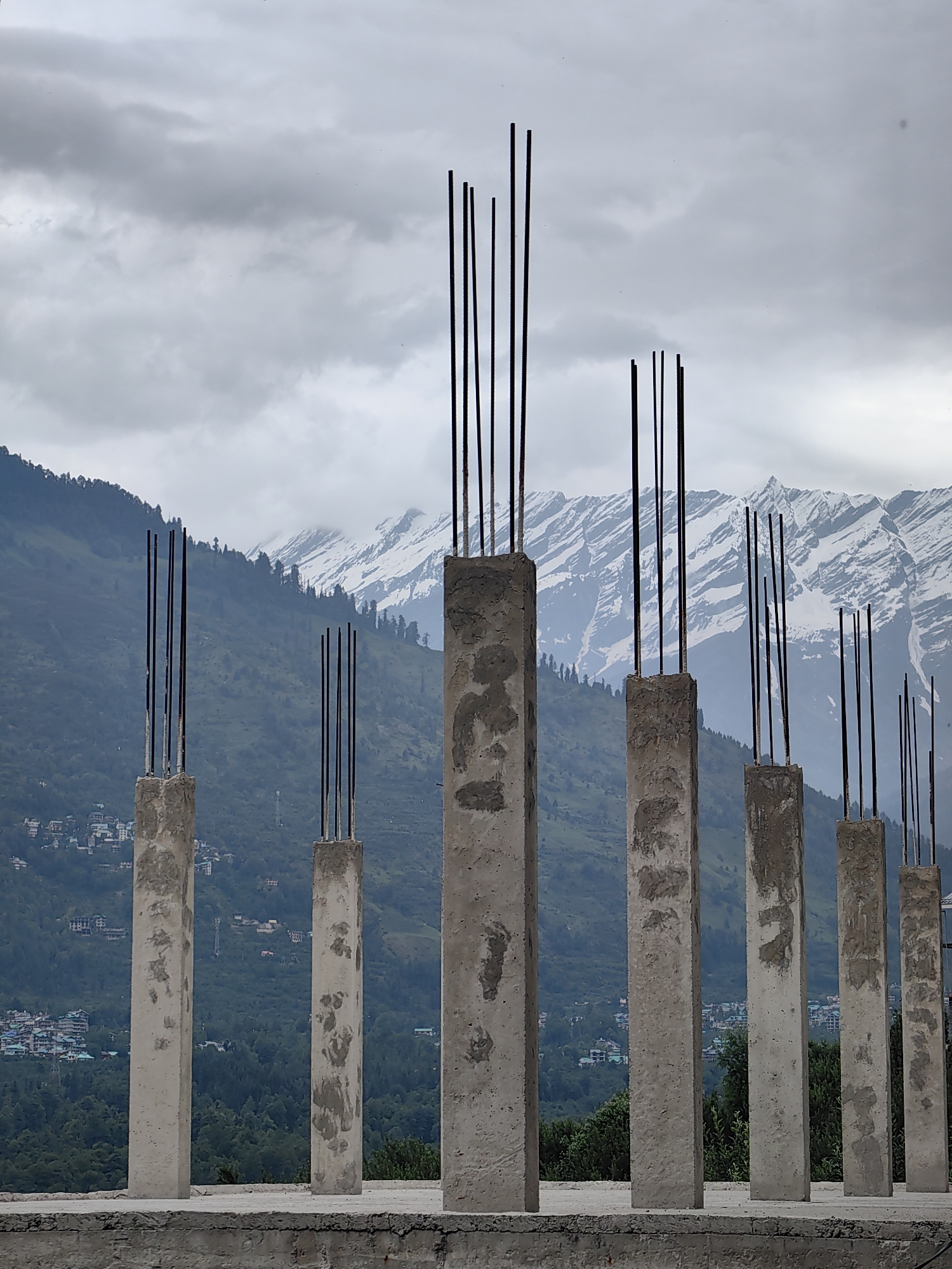 The image shows a series of unfinished concrete pillars with rebar protruding from their tops. They are set against a backdrop of a lush green valley with a small town nestled on the hillside, and snow-capped mountains in the distance. The sky is overcast with heavy clouds. This scene is characteristic of a construction site in the town of Vashisht, India, known for its scenic views and mountainous terrain.