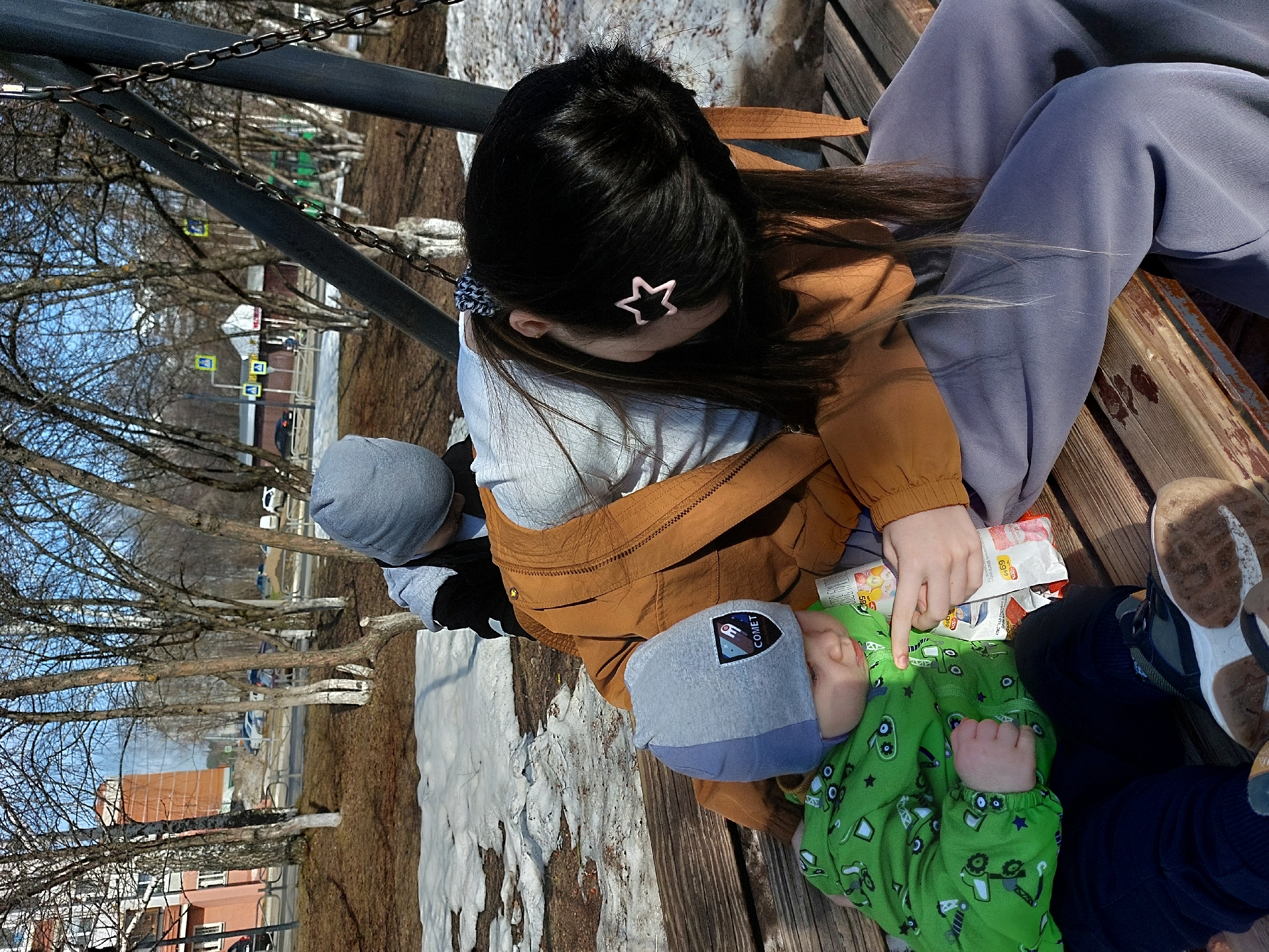 A woman and two children are sitting on a wooden bench outdoors. The woman has dark hair with a star-shaped clip and is wearing a grey shirt and brown jacket, as well as light purple sweatpants. One child, dressed in a green patterned onesie and a two-toned grey and blue hat, is lying back on the bench. The other child is wearing a grey hat, a black and white jacket, and dark blue pants. The woman's hand is touching a packet of chips next to the child in the green onesie. The bench is positioned in an area with melting snow and bare trees, suggesting it is early spring. In the background, there are buildings and street signs visible, indicating an urban location. Based on visual cues such as architecture and street signage, the location is in Chaikovsky, Russia. A swing structure with metal chains is also visible near the bench.