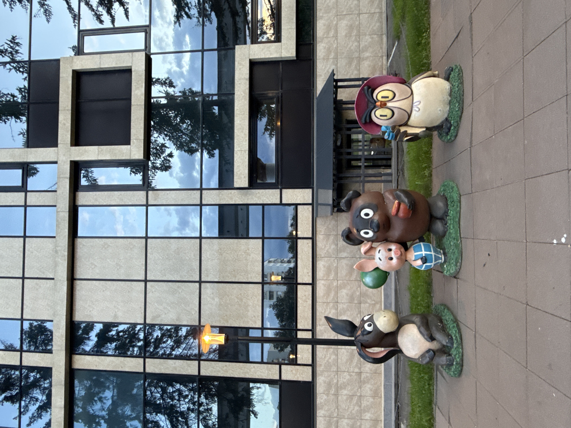 The image, oriented vertically, captures a scene featuring four cartoon character statues arranged on a paved sidewalk, set against the backdrop of a modern building. To the left, the multi-story building displays a facade of large glass windows interspersed with light-colored stone or tile panels, forming a grid pattern. The glass reflects parts of a cloudy sky and the dark branches of trees.
On the right side of the image, from top to bottom, are the four statues. The uppermost statue is an owl, cream-colored with brown wings and yellow eyes, wearing a red hat and a light blue bow tie. Below the owl is a dark brown bear with a round black nose and white eyes, holding a small brown object. Nestled next to the bear is a pink piglet wearing a blue and white checkered shirt and holding a green balloon. The last statue, at the bottom, is a dark gray/black donkey with a lighter face and belly, a white patch on its nose, and long ears. All statues are standing on small green bases resembling grassy patches.
A street light with a glowing, warm-toned lamp is visible on the right, positioned behind the donkey statue, suggesting it might be dusk or early evening. The scene is static, with no human activity present. There is no visible text. The ground consists of gray rectangular pavers, with narrow strips of green grass running alongside.