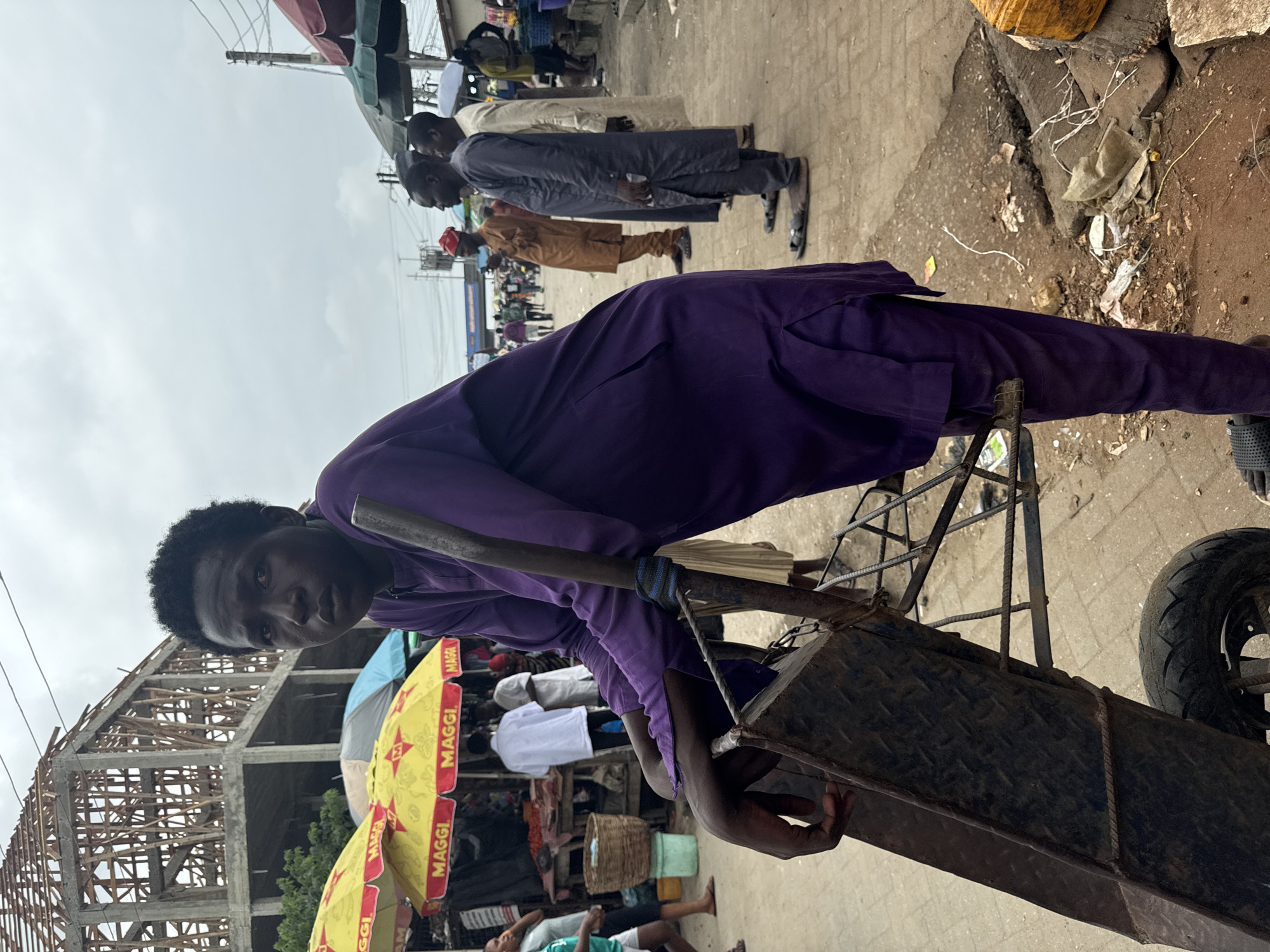 In Ado, Nigeria, a young man in a purple tunic and trousers stands next to a metal wheelbarrow, leaning on its handle. He has dark skin and short, dark hair. The wheelbarrow is worn and rusty, with a metal tray and wire basket. In the background, a building under construction with a wooden frame is visible. A market scene unfolds with several people browsing stalls under colorful umbrellas, one of which is branded with "MAGGI". The ground is a mix of dirt and paved surfaces. The sky is overcast.