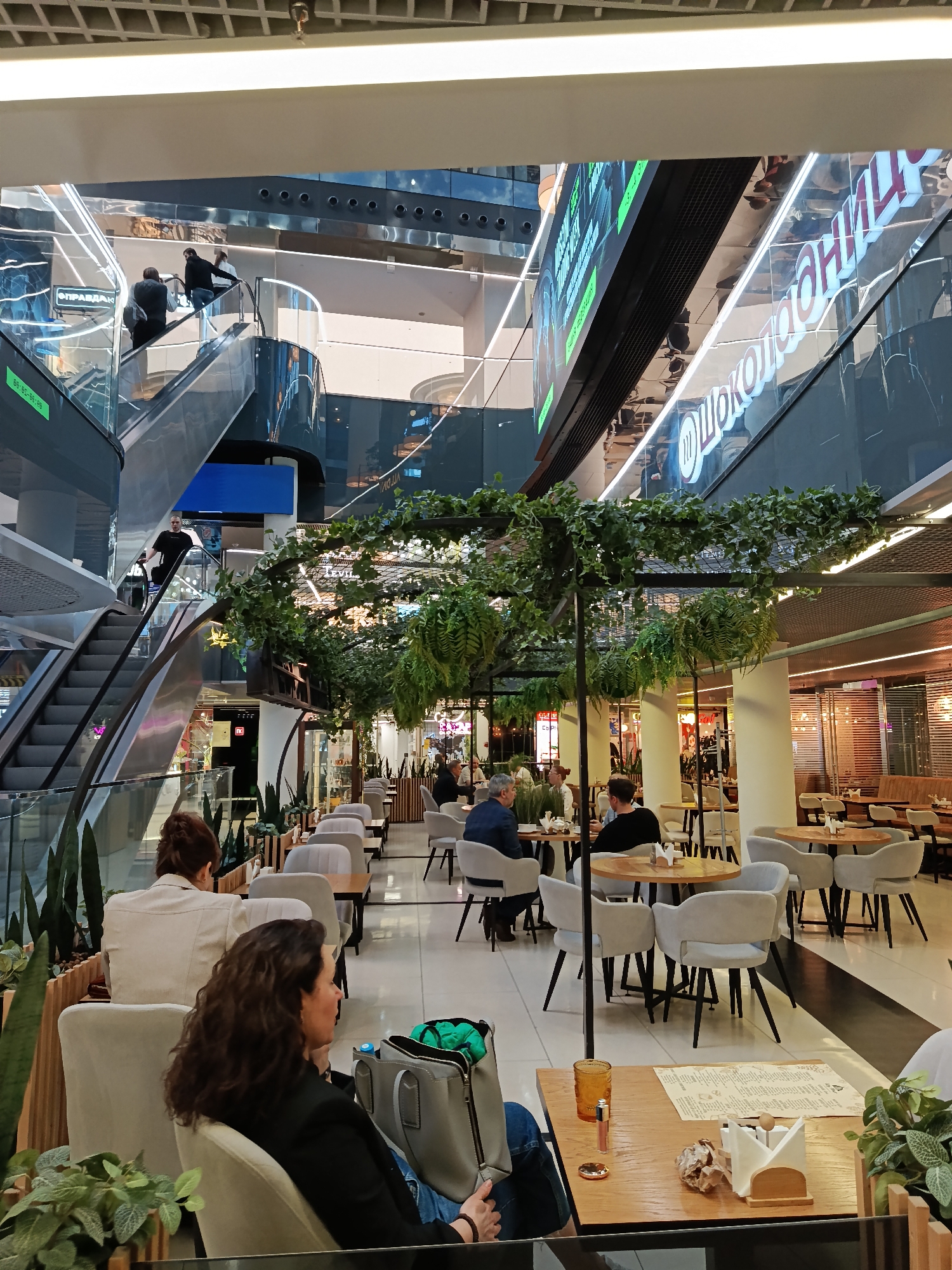 The image captures the interior of a multi-story shopping mall in Moscow, Russia. The space includes a seating area that seems to be part of a restaurant or cafe. People are seated at tables, some are in conversation, while others appear to be waiting. The seating area is decorated with greenery hanging from overhead structures. In the background, escalators are visible with people riding them between floors. Reflected signage in Russian can be seen on the upper levels of the mall.