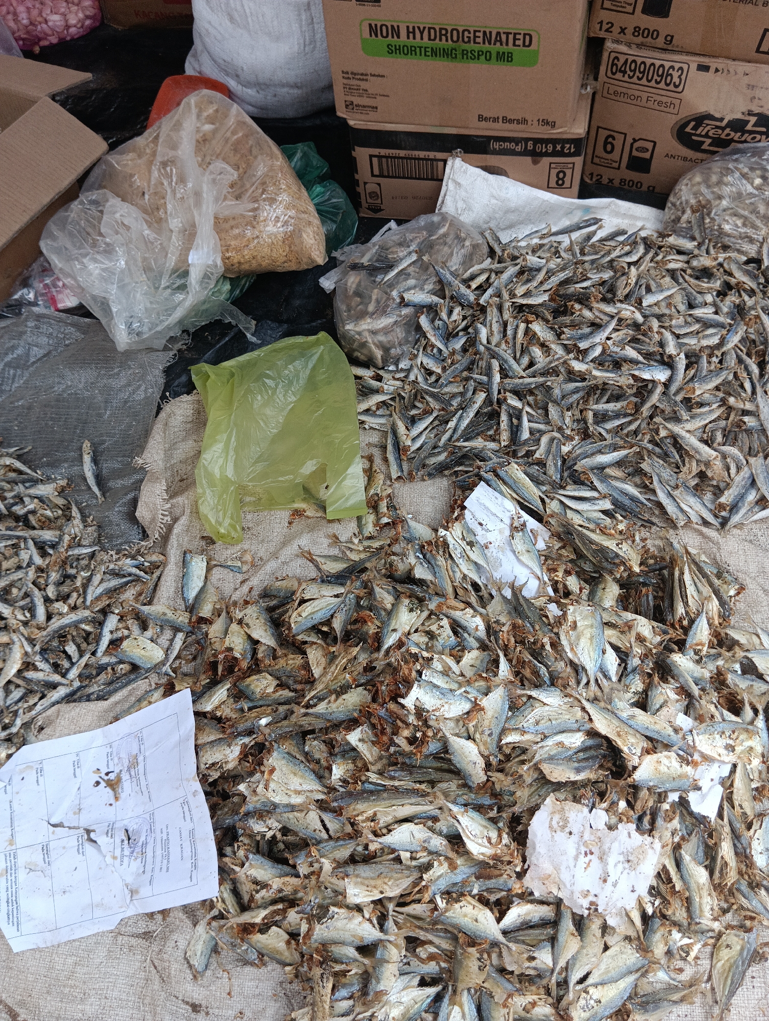 The image depicts a market stall in Binakarsa, Indonesia, featuring a large pile of dried fish. The fish are small and appear to be sardines or similar species, laid out on a rough, woven mat. Interspersed with the fish are several plastic bags, one bright yellow and others clear, containing what looks like grains or other food items. In the background, cardboard boxes are stacked, with labels indicating products such as "NON HYDROGENATED SHORTENING RSPO MB" and "Lifebuoy ANTIBAC". A white piece of paper, possibly a receipt or label, lies on top of some of the fish. The overall scene suggests a food market environment with goods for sale.