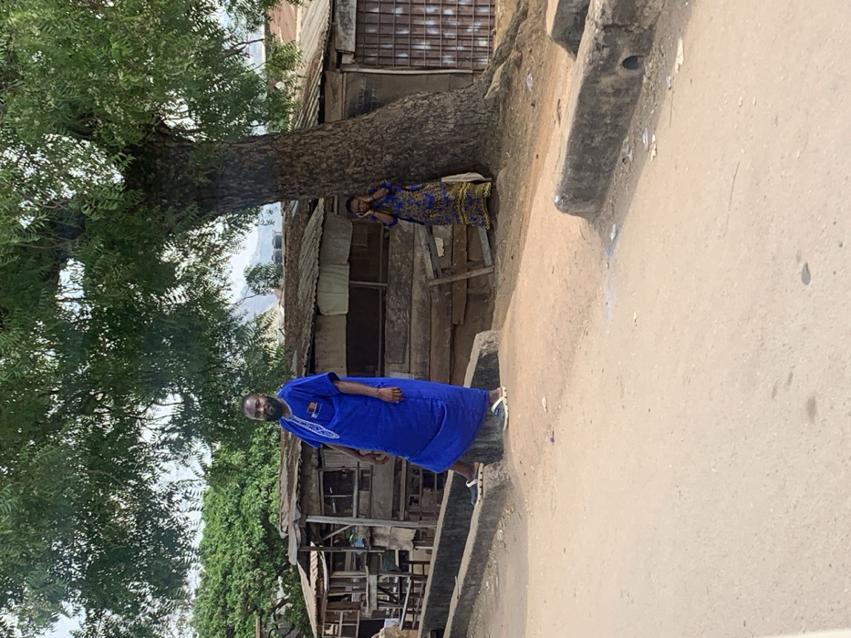 This image depicts an outdoor urban scene in the city of Egbedi, Nigeria. In the foreground, a light brown dirt road spans horizontally, interrupted by two parallel, low concrete structures resembling curbs or drainage channels. The left concrete structure is partially obscured by the subject on the left, while the right structure shows a dark, circular opening on its face.
Two individuals are present. On the left, a dark-skinned adult male with a full beard stands facing the camera. He wears a long, loose-fitting royal blue traditional garment (likely a kaftan or agbada) with white embroidery detailing around the neck and chest. A small, multi-colored emblem, possibly a flag, is visible on his left chest pocket. He wears dark sandals or clogs with prominent white soles; his left foot rests on the far concrete structure, while his right foot is on the dirt road next to it.
To the right, a dark-skinned adult female stands partially concealed behind the trunk of a large tree with a textured bark and visible surface roots. She wears a long, patterned dress, predominantly blue with yellow and black motifs. Her hands are raised to the sides of her head, an action possibly associated with holding a phone or reacting to an auditory stimulus.
Behind the individuals and the tree, a series of rudimentary structures, likely shops or dwellings, are visible. These buildings are constructed from weathered wooden planks and feature corrugated metal roofs, some with evidence of rust. Lush green foliage from various trees forms a dense canopy above the scene and extends into the background, where distant, undulating hills are discernable under an overcast sky.