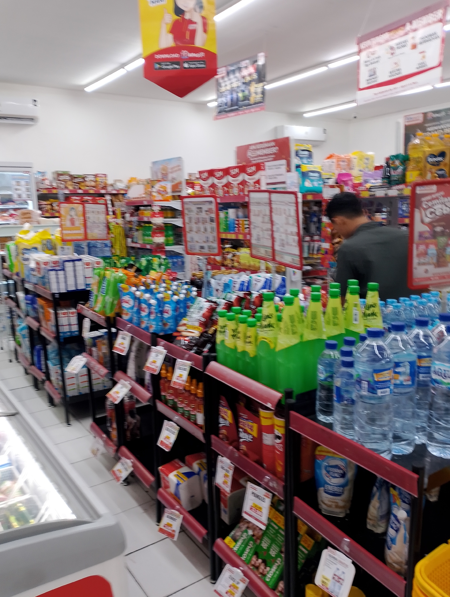 The image depicts the interior of a brightly lit convenience store, likely an Alfamart branch, in Magelang, Indonesia. Shelves are stocked with a variety of products, primarily beverages and packaged snacks. In the foreground, numerous bottles of water are prominently displayed, along with brightly colored bottles of what appear to be soft drinks or juices with green caps. Behind these, various packaged food items, including chips and other snacks, are visible. A man, wearing a dark green shirt, is seen from the back, partially obscured by the product displays, and appears to be browsing or selecting items. Advertising banners are hung from the ceiling, featuring cartoon characters and promotional messages. The store environment is clean and organized, with tiled flooring and visible lighting fixtures. A refrigerated display case is partially visible on the left side of the image.