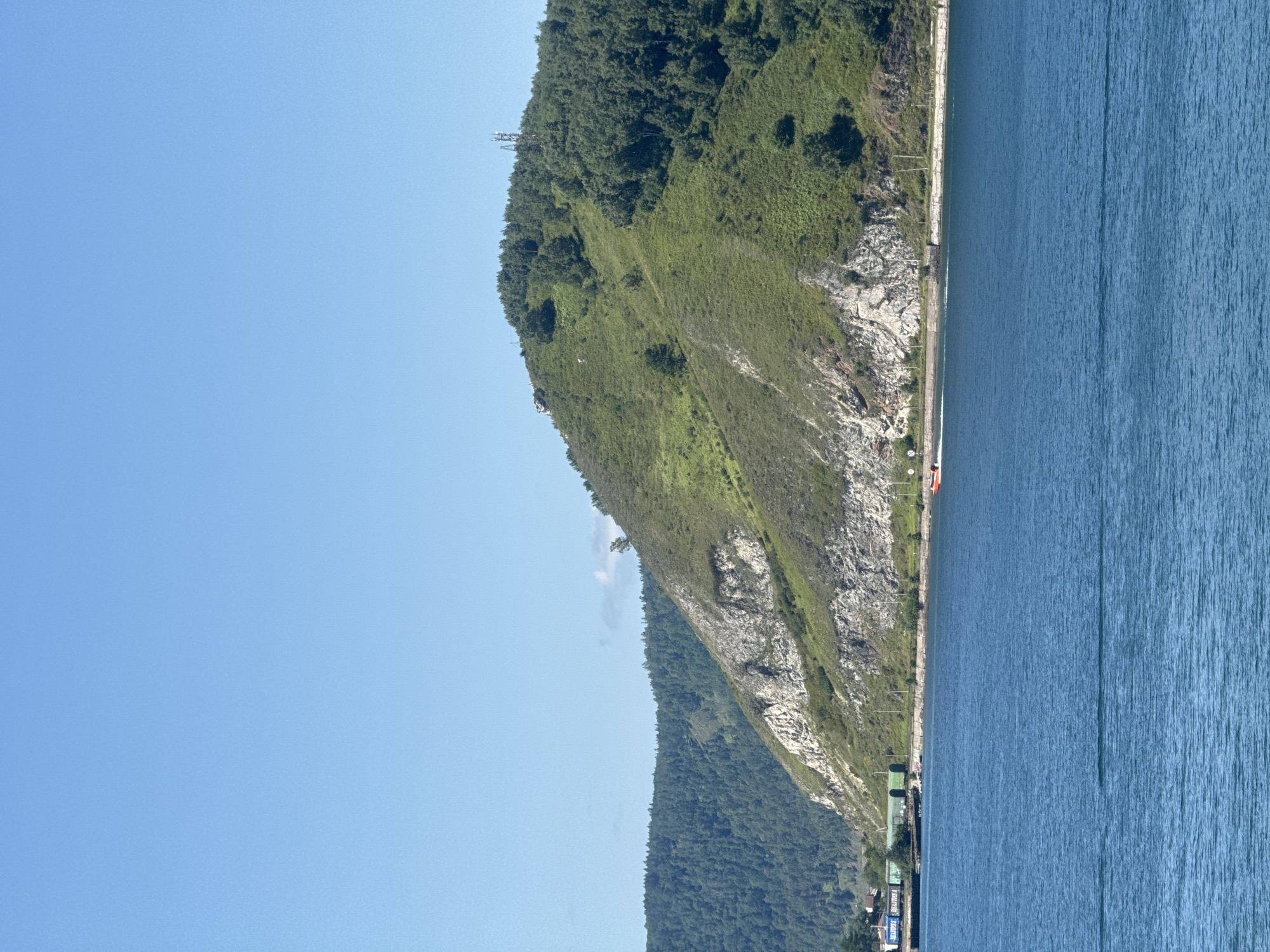 A wide shot captures a bright, sunny day on a body of water with a steep, green hillside rising behind it. The water is a deep blue and calm, with subtle ripples. Along the base of the hillside, a road and a railway line are visible. A train with green carriages is stopped on the tracks, and a small orange boat is moving on the water near the shore. The hillside is covered in lush green grass and trees, with rocky outcrops and a few solitary trees scattered across its surface. A communications tower stands on the summit of the hill. In the background, more forested hills recede into the distance under a clear blue sky. This scene is characteristic of the landscape found in Listvyanka, Russia, a village located on the southwestern shore of Lake Baikal.