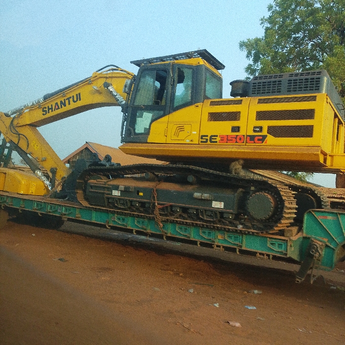 A yellow Shantui SE350LC excavator rests on a green flatbed trailer. The excavator’s boom arm extends to the left, with the cabin and machinery body centered on the trailer. The trailer is secured with chains. The ground appears to be dirt or clay. A tree is visible in the background on the right, and the sky is overcast.